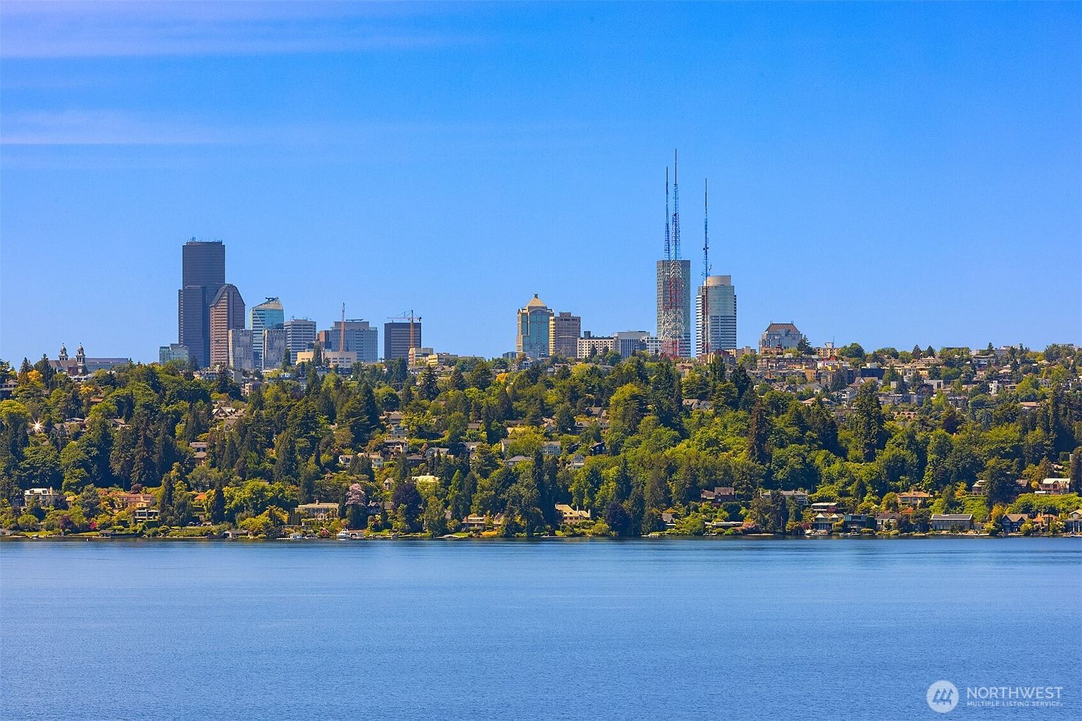 This wide-angle, elevated shot captures a stunning panoramic view of the Seattle skyline rising above a lush, tree-covered hillside and the calm waters of Lake Washington. The composition highlights the contrast between the dense urban architecture and the natural beauty of the surrounding landscape, emphasizing the property's desirable location. The clear blue sky and bright daylight create an inviting and aspirational atmosphere, perfect for showcasing a premium real estate setting.