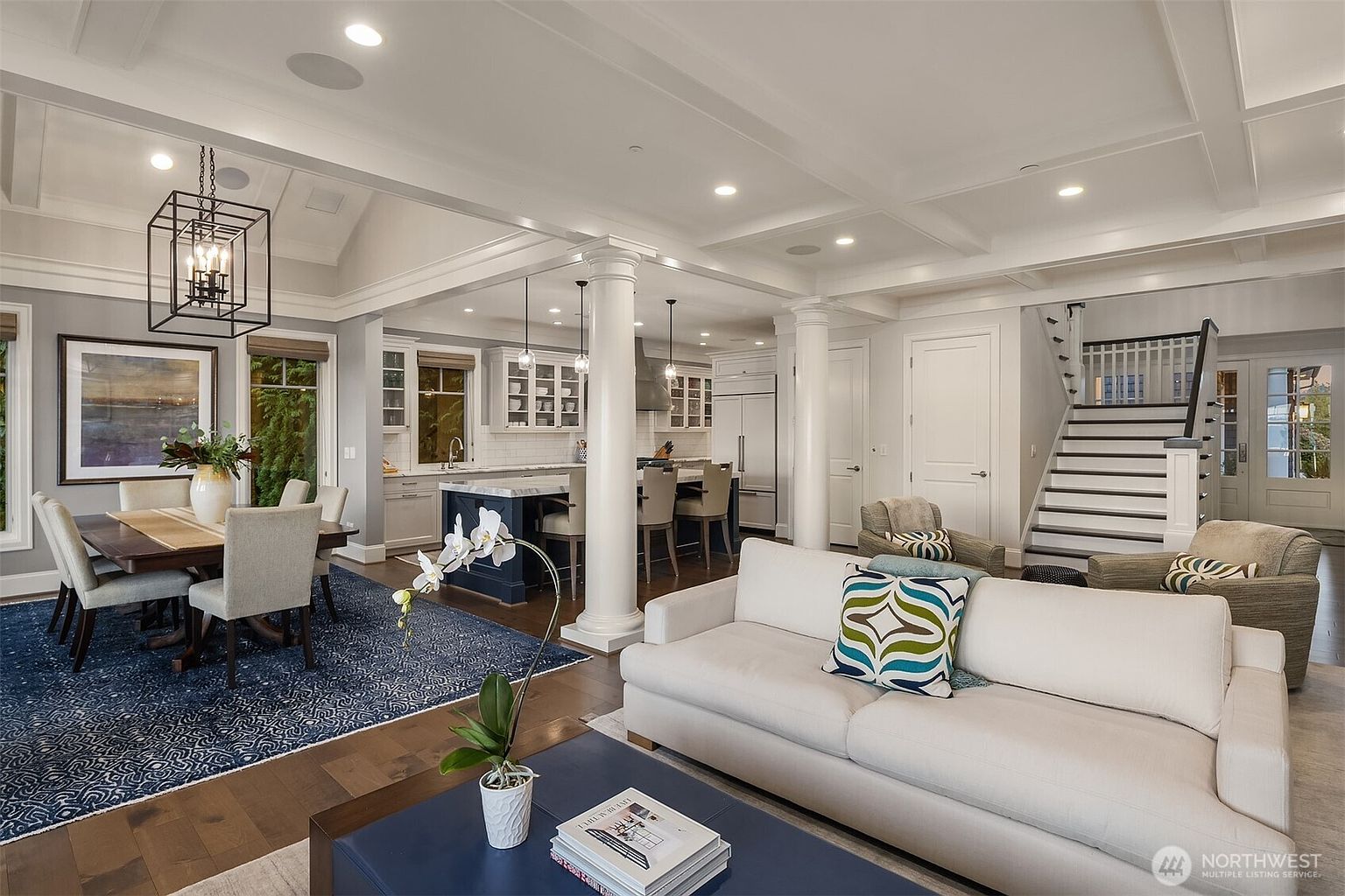 This open-concept living space features a seamless transition between a formal dining area, a gourmet kitchen, and a comfortable living room. The room is characterized by high coffered ceilings, elegant white columns, and a sophisticated color palette of navy blue and neutral tones. The perspective captures the airy, expansive layout, highlighting the high-end finishes and the inviting, well-lit atmosphere perfect for modern family living.