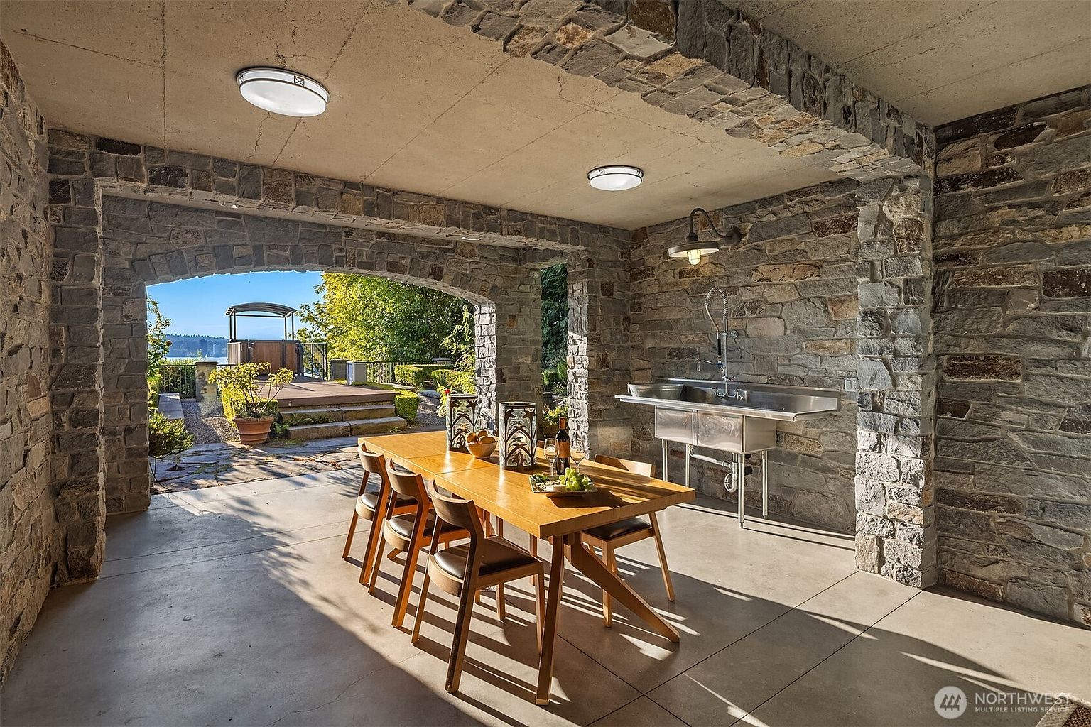 This covered outdoor dining area features rustic stone walls and arches, creating a sophisticated and durable space for al fresco entertaining. The area is furnished with a warm-toned wooden dining table and chairs, complemented by a stainless steel utility sink for convenience. The open-air design frames a picturesque view of the lush backyard and distant water, offering a seamless transition between indoor comfort and outdoor beauty.