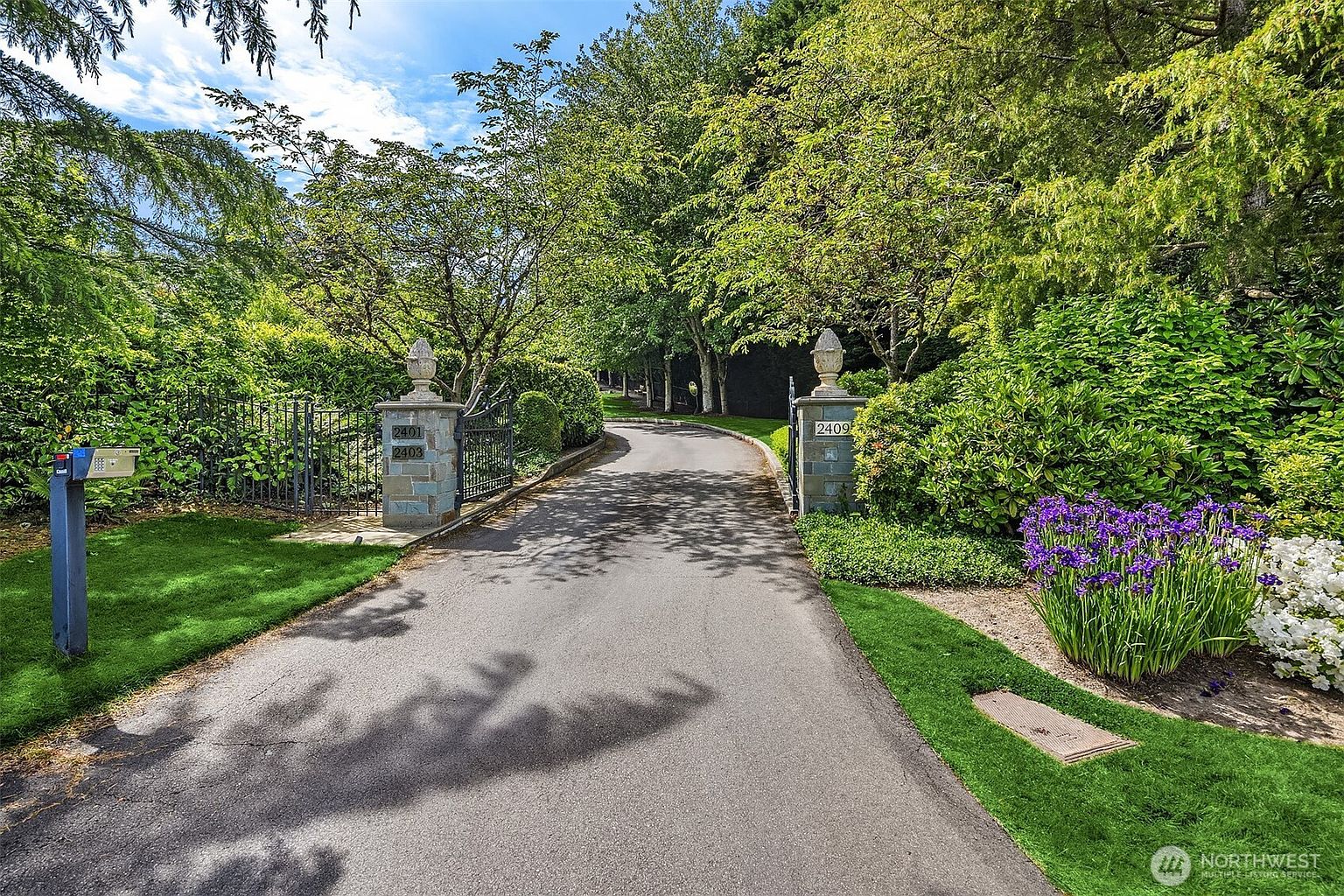 This image captures a grand, gated driveway entrance leading to a private estate, framed by lush, mature trees and manicured landscaping. The stone pillars, topped with decorative finials, feature address numbers 2401, 2403, and 2409, creating an inviting and prestigious first impression. The perspective is a straight-on view from the street, emphasizing the depth and seclusion of the property.
