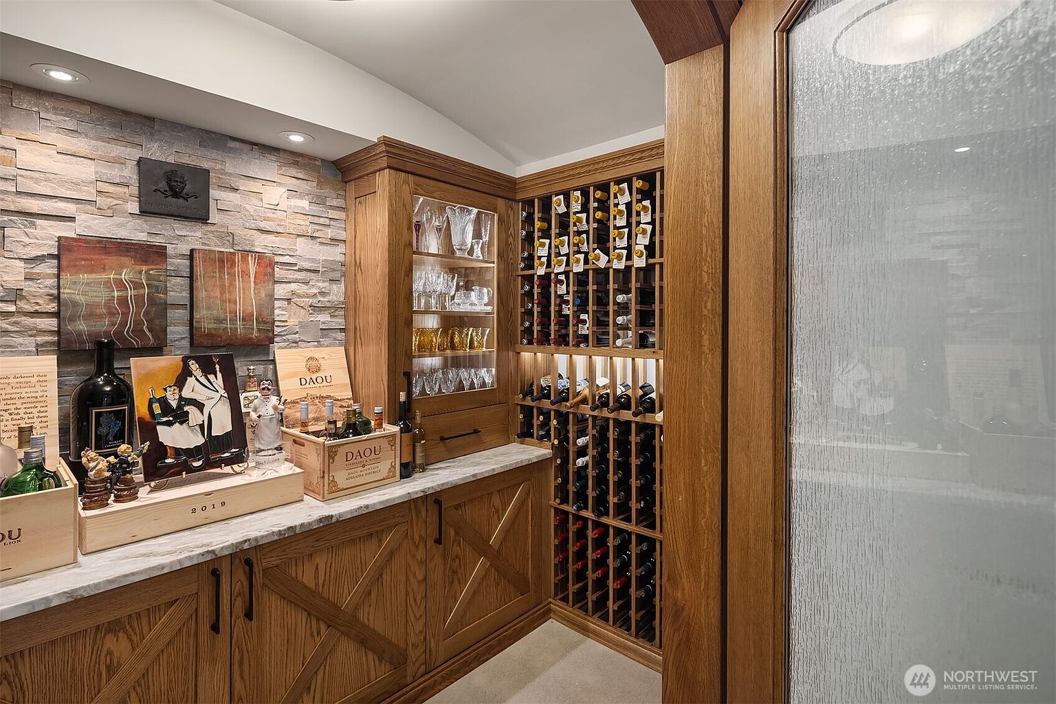This sophisticated wine cellar features custom oak cabinetry, a stone-tiled accent wall, and a dedicated glass-fronted display cabinet for glassware. The space is elegantly appointed with marble countertops, wine storage racks, and decorative art pieces, creating a refined and inviting atmosphere for wine enthusiasts. The perspective is a medium shot capturing the warmth of the wood tones and the organized storage layout.