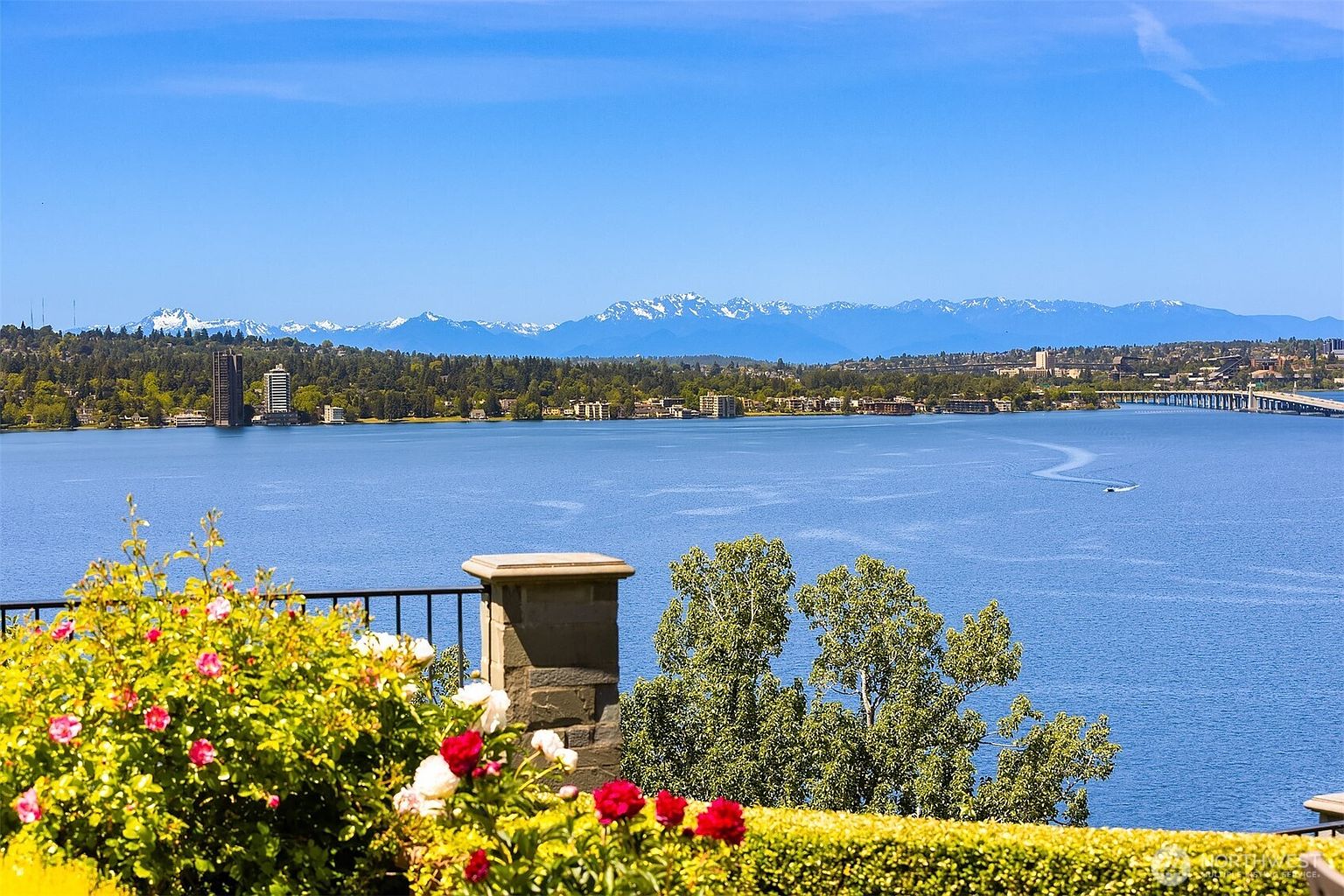 This image captures a breathtaking, elevated view from a balcony overlooking a vast, serene lake with a distant bridge and snow-capped mountain range. In the foreground, a vibrant rose bush with blooming red and white flowers adds a touch of natural elegance, framing the expansive water vista. The perspective is cinematic and tranquil, emphasizing the property's premium waterfront location and scenic landscape.
