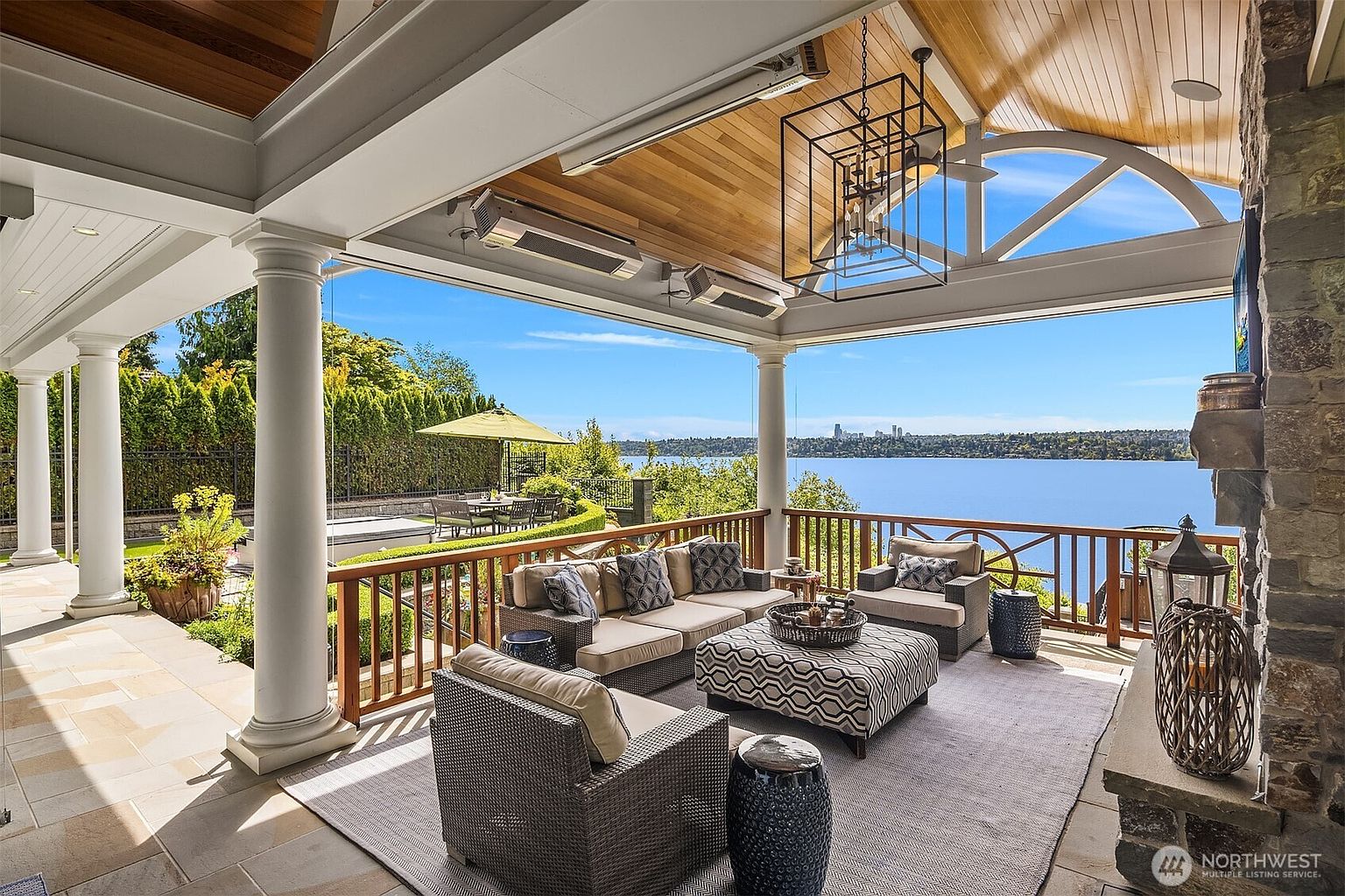 This luxurious covered patio offers a stunning panoramic view of a lake and distant city skyline, creating an ideal space for outdoor relaxation and entertaining. The area is furnished with comfortable, high-end wicker seating, a patterned ottoman, and a stone fireplace, all set under a beautiful wood-paneled ceiling with integrated heaters and a large chandelier. The perspective captures the seamless transition between the elegant outdoor living space and the serene waterfront landscape.