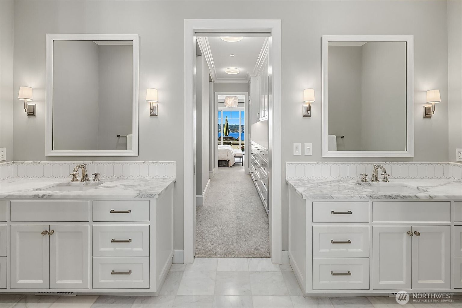 This elegant primary bathroom features a symmetrical double vanity with white cabinetry, marble countertops, and matching rectangular mirrors flanked by wall sconces. A central doorway leads into a long, carpeted walk-in closet that provides a view through to a bright bedroom and outdoor scenery. The space is characterized by a clean, sophisticated aesthetic with high-end finishes and a bright, airy atmosphere.