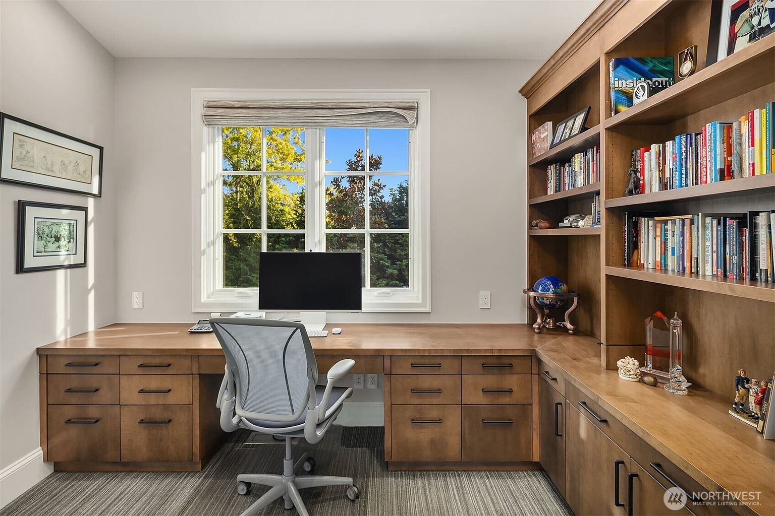 This sophisticated home office features custom built-in wooden cabinetry and shelving that wraps around the corner, providing ample storage and display space. A central desk area is positioned beneath a large window that offers a bright, natural view, complemented by a modern ergonomic office chair. The room is finished with neutral walls, framed artwork, and a textured carpet, creating a professional and inviting workspace.