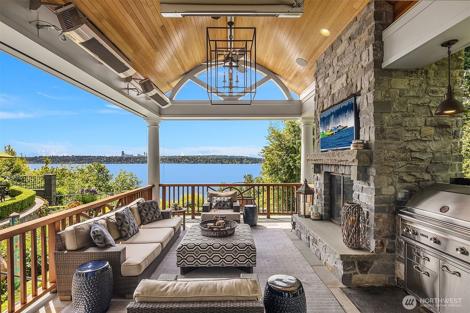 This luxurious covered outdoor living space features a stone fireplace with an integrated television, a built-in stainless steel grill, and comfortable lounge seating. The vaulted wood-paneled ceiling and expansive open archway frame a breathtaking, unobstructed view of the lake and distant city skyline. The space blends rustic stone elements with refined, modern furnishings, creating an ideal setting for high-end outdoor entertaining.