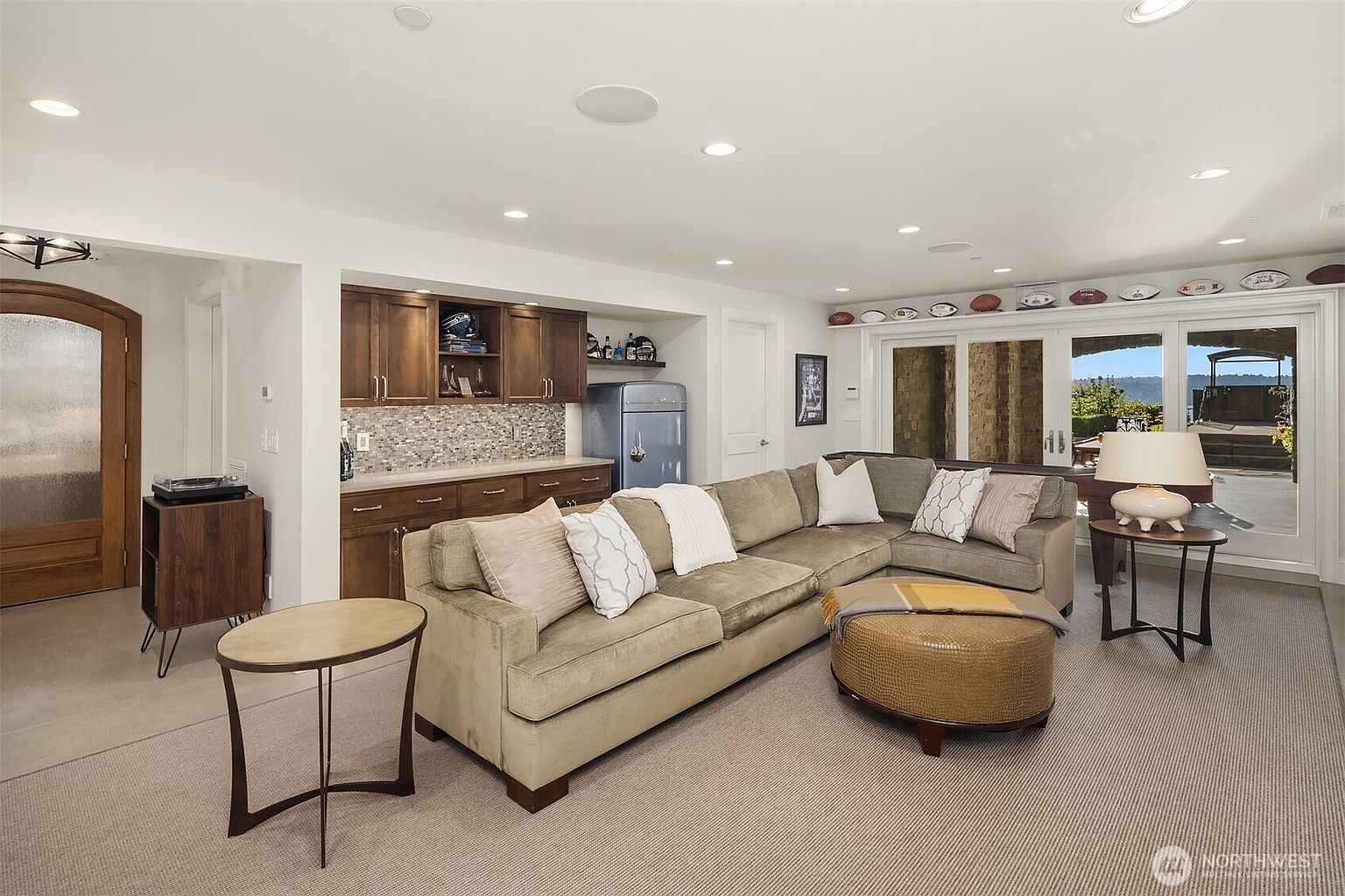 This inviting living room features a large, plush sectional sofa paired with a circular ottoman, creating a comfortable space for relaxation. A built-in wet bar with dark wood cabinetry and a retro-style blue refrigerator adds a functional and stylish touch, while large glass doors provide a seamless transition to the outdoor area. The room is well-lit with recessed lighting and decorated with a collection of sports memorabilia, offering a warm and casual atmosphere.
