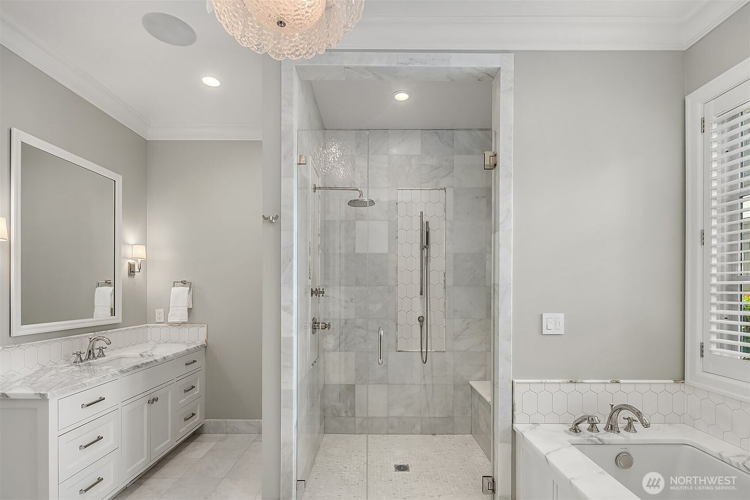 This luxurious primary bathroom features a spacious walk-in shower with marble tile, a built-in bench, and a rainfall showerhead. To the left, a white vanity with marble countertops and dual sinks provides ample storage, while a deep soaking tub is positioned to the right. The room is bathed in soft, neutral tones, creating an elegant and serene spa-like atmosphere.