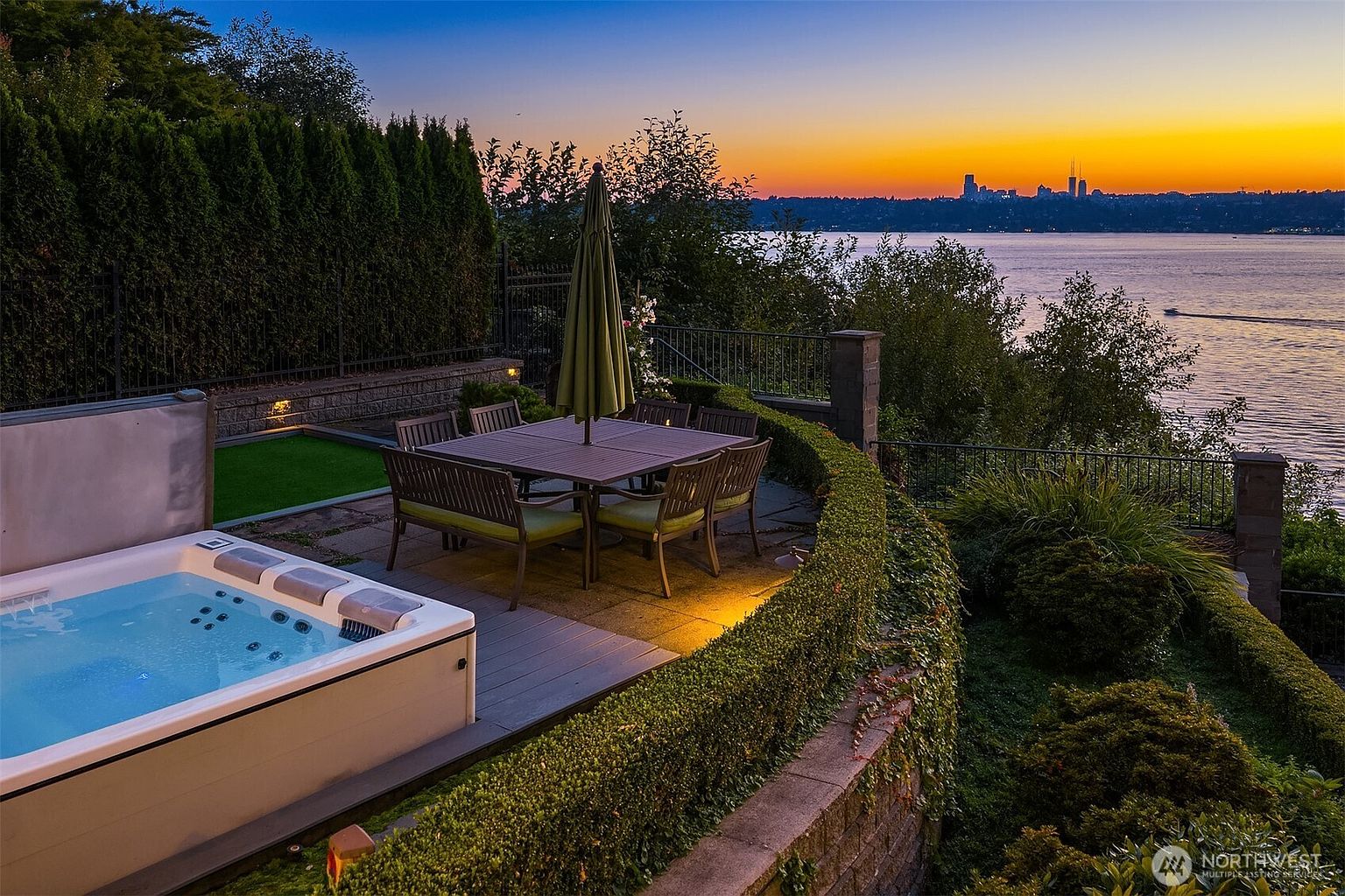 This stunning outdoor terrace features a luxurious hot tub and a dining set, perfectly positioned to capture a breathtaking sunset view over the water and distant city skyline. The space is framed by manicured hedges and stone retaining walls, creating an intimate and sophisticated atmosphere for evening relaxation. The perspective is elevated, emphasizing the seamless blend of private luxury and expansive natural beauty.