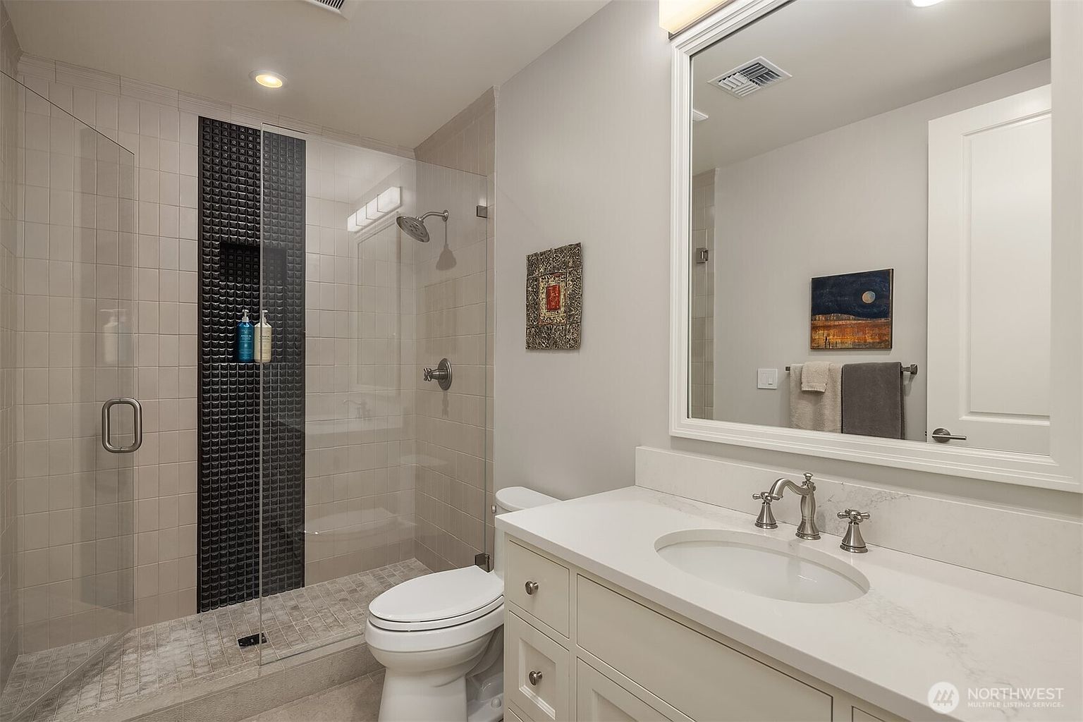 This modern bathroom features a sleek walk-in shower with a striking black tiled accent wall and a glass enclosure. The vanity is equipped with a white countertop, a classic undermount sink, and a large, framed mirror that enhances the sense of space. The neutral color palette and clean lines create a bright, contemporary, and inviting atmosphere.