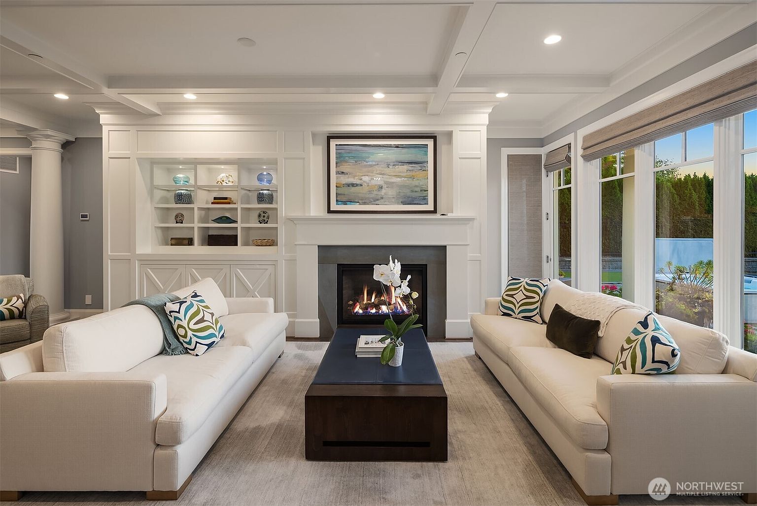 This elegant living room features a symmetrical layout with two plush white sofas facing each other across a dark wood coffee table. A central fireplace with a stone surround and a large abstract painting serves as the focal point, flanked by built-in white shelving units. The room is bathed in natural light from large windows, creating a bright, sophisticated, and inviting atmosphere.