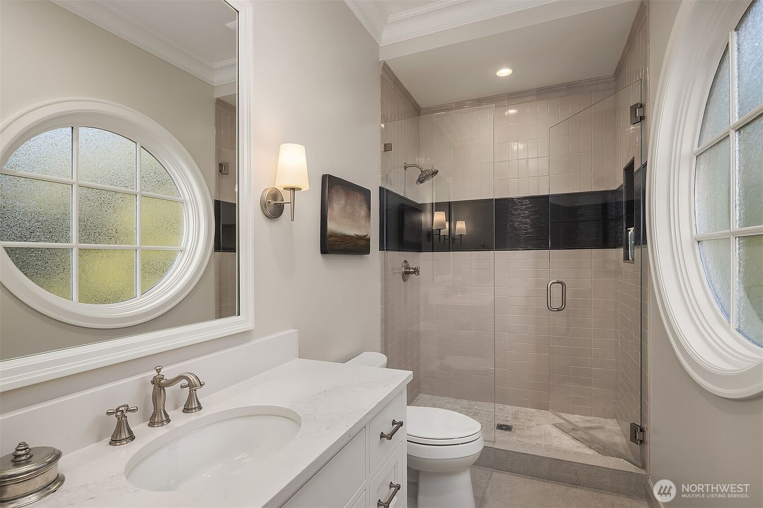 This elegant bathroom features a clean, contemporary aesthetic with a white vanity topped by a light-colored stone countertop and a polished chrome faucet. A glass-enclosed walk-in shower with a striking black tile accent band adds a modern touch, while a unique circular window allows natural light to brighten the space. The neutral color palette and thoughtful lighting create a sophisticated and inviting atmosphere.