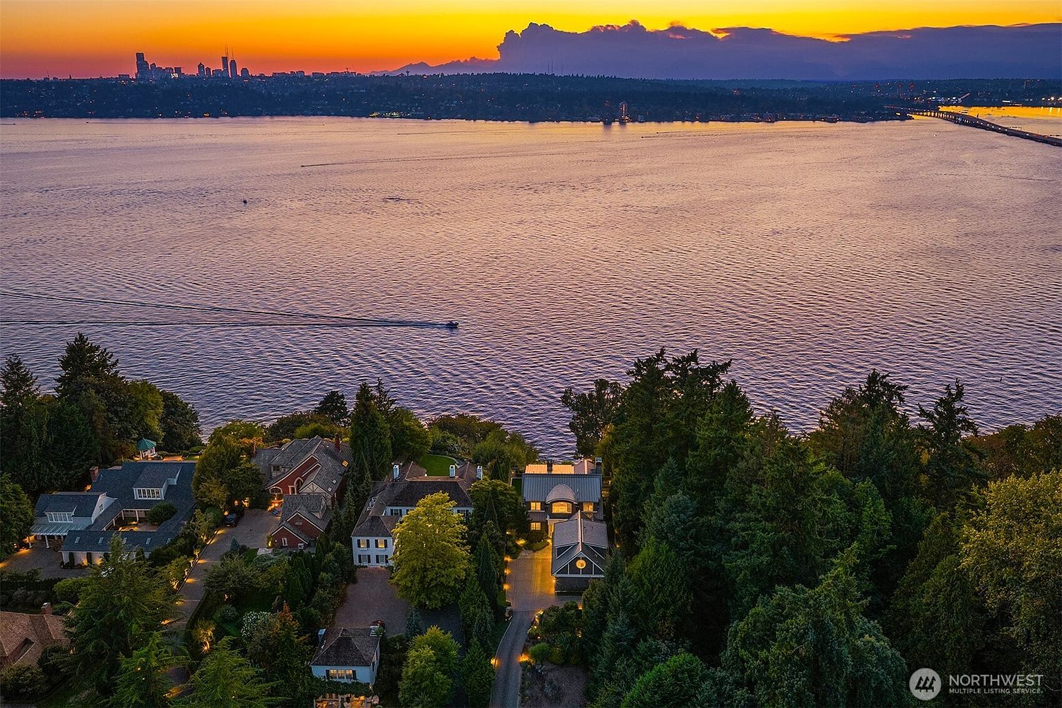 This stunning aerial view captures a luxury waterfront property at sunset, overlooking a vast lake with a distant city skyline. The scene highlights the home's secluded, tree-lined setting, featuring a multi-gabled roofline and warm, inviting exterior lighting that contrasts beautifully with the deep blue twilight. The perspective emphasizes the expansive water views and the peaceful, high-end residential atmosphere of the neighborhood.