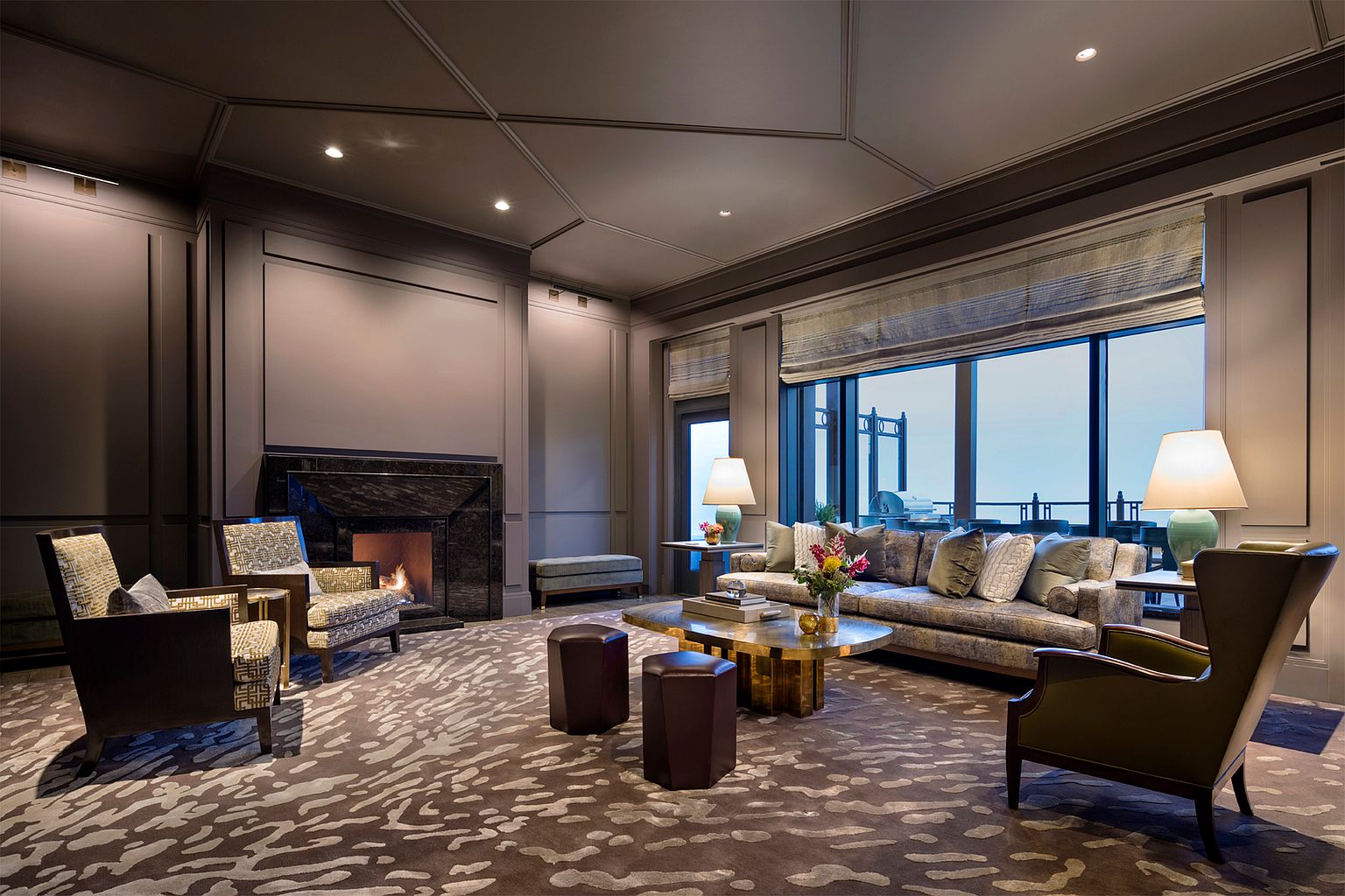 This sophisticated living room features a dark, moody color palette with paneled walls and a striking patterned carpet. A central seating area is anchored by a gold-toned coffee table, flanked by a plush sofa, two patterned armchairs, and a leather wingback chair. A dark stone fireplace surround serves as a focal point, while large windows with Roman shades provide a view of the outdoor balcony.