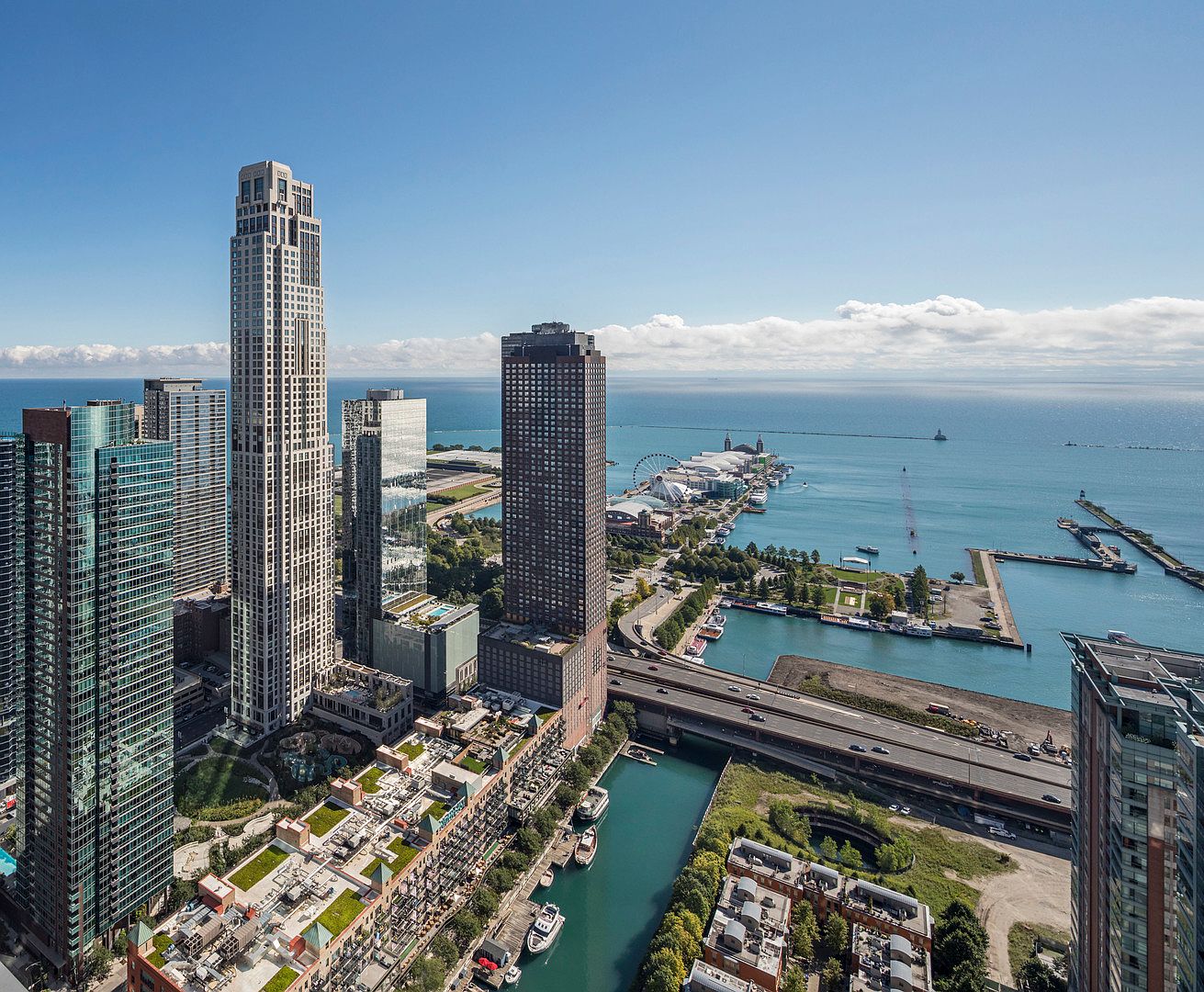 This high-altitude aerial view captures a dense urban skyline featuring several prominent glass-clad skyscrapers situated along a waterfront. The perspective highlights the proximity to a large body of water, a marina with docked boats, and a nearby amusement pier, showcasing a vibrant metropolitan lifestyle. The scene is bathed in bright daylight, emphasizing the modern architectural density and the strategic location of these high-rise residences.