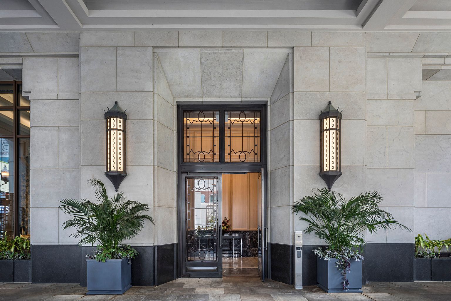This elegant building entrance features a symmetrical design with light-toned stone cladding and a dark, ornate metal-framed glass door. Two tall, decorative wall-mounted lanterns flank the doorway, while large potted plants add a touch of greenery to the formal stone-paved landing. The perspective is a straight-on, eye-level shot that emphasizes the sophisticated and welcoming architectural details of the property.