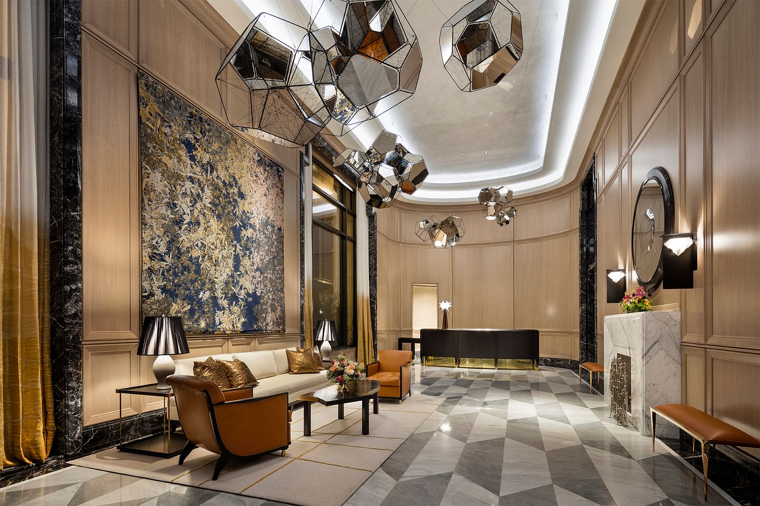 This sophisticated lobby or lounge area features a high-end, contemporary design with light wood paneling and a striking geometric patterned stone floor. The space is anchored by a plush seating area with leather armchairs and a sofa, complemented by a marble-clad fireplace surround and dramatic, sculptural geometric light fixtures. The perspective is a wide-angle shot looking toward a reception desk, emphasizing the grand scale and luxurious atmosphere of the building's common area.