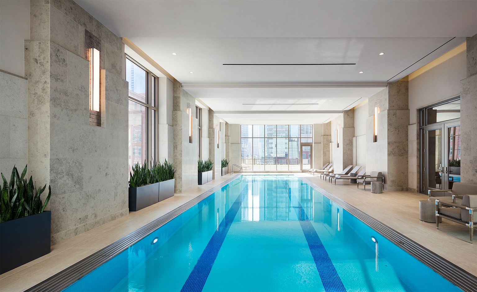This indoor lap pool area features a sleek, minimalist design with light-toned stone walls and large windows that provide an expansive view of the city skyline. The space is accented by potted snake plants and modern lounge chairs arranged along the side, creating a serene and upscale atmosphere. The perspective is centered down the length of the pool, emphasizing the clean lines and bright, airy aesthetic of the facility.