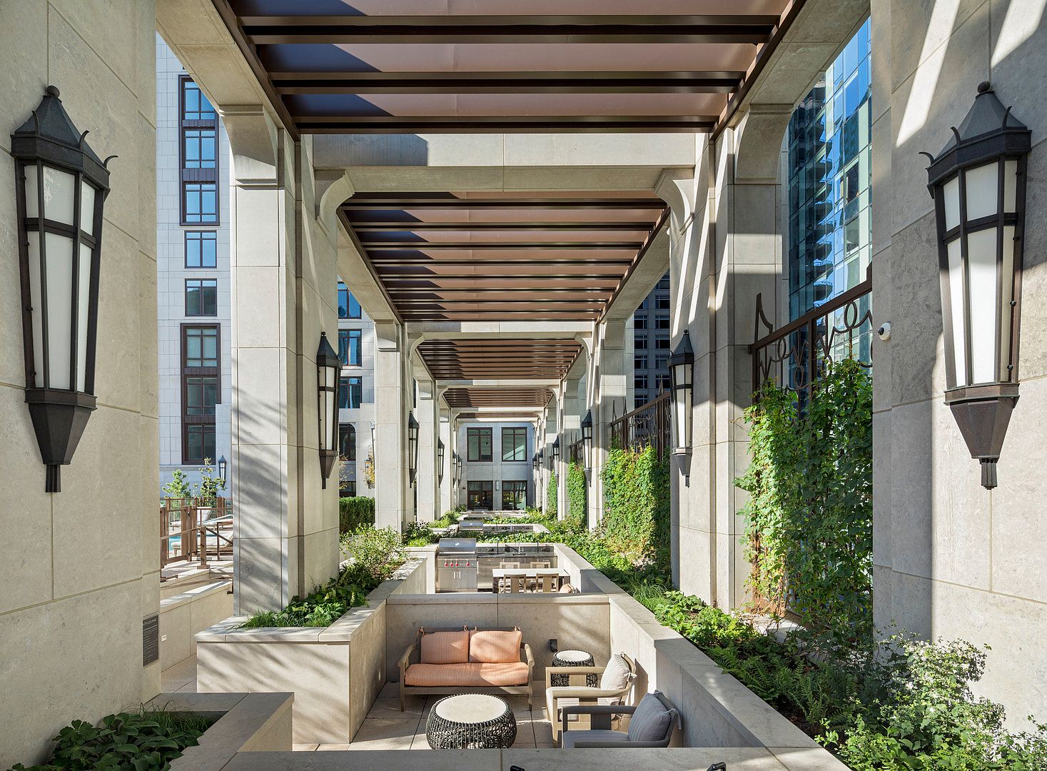 This sophisticated outdoor lounge area features a covered pergola structure with stone pillars and elegant wall-mounted lanterns. The space includes a sunken seating area with plush furniture and a nearby outdoor kitchen, all surrounded by lush greenery and climbing vines. The perspective looks down the length of the corridor, highlighting the architectural symmetry and upscale design of this communal terrace.