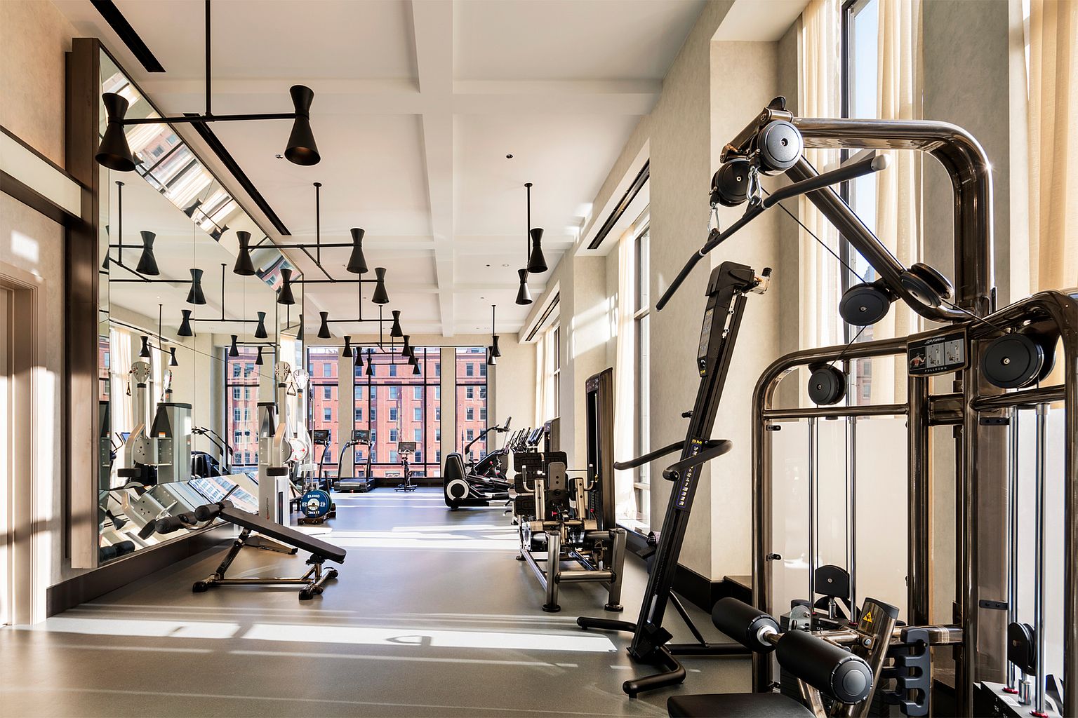 This bright and spacious home gym features a variety of professional-grade fitness equipment, including a cable machine, a weight bench, and cardio machines. The room is characterized by a long wall of mirrors that enhances the sense of space, complemented by tall windows that allow natural light to flood the area. The clean, modern aesthetic is accented by unique, multi-armed pendant lighting fixtures suspended from the ceiling.