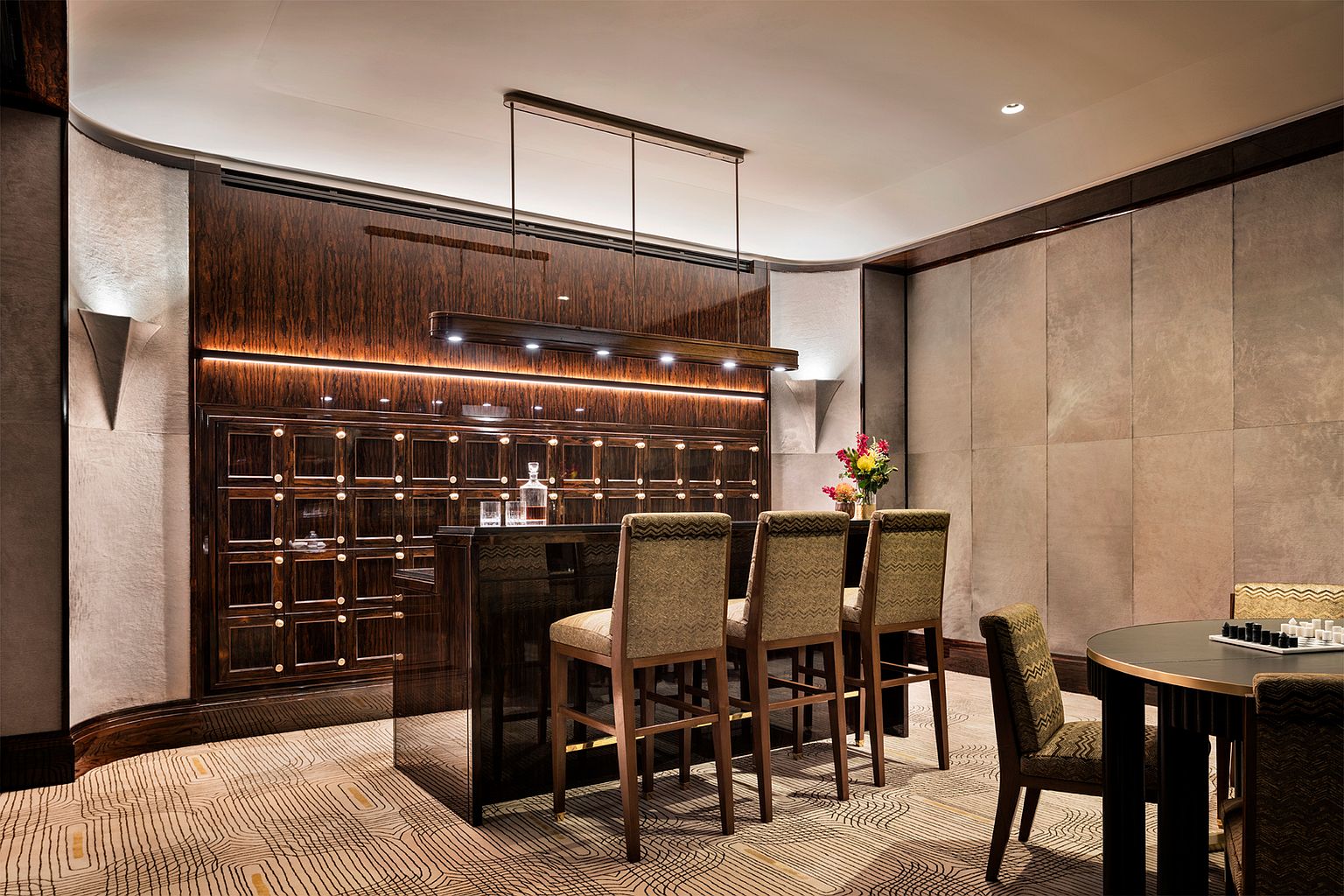 This sophisticated lounge area features a dark, polished wood bar with integrated storage lockers and a linear pendant light fixture. Three upholstered bar stools are positioned at the counter, while a small round table with a chessboard sits in the foreground. The room is accented by textured wall panels, warm under-cabinet lighting, and a patterned carpet, creating an upscale, club-like atmosphere.