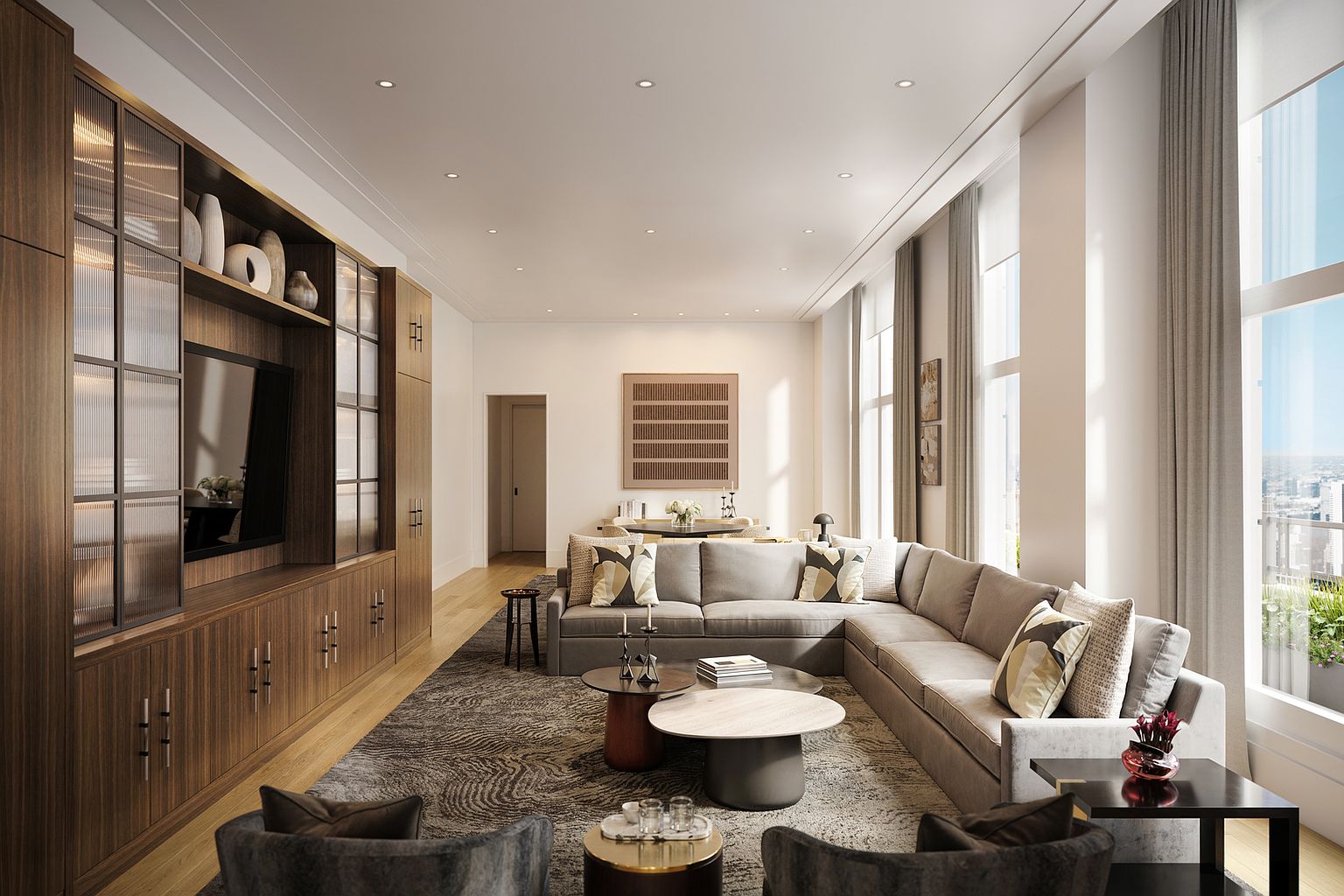 This sophisticated living room features a large, L-shaped sectional sofa arranged on a textured area rug, centered around a pair of nested coffee tables. A custom wood-paneled wall unit with integrated shelving and a television provides a focal point, while tall windows along the right wall invite abundant natural light and offer city views. The space is decorated in a neutral, contemporary palette with elegant lighting and a seamless transition to a dining area in the background.