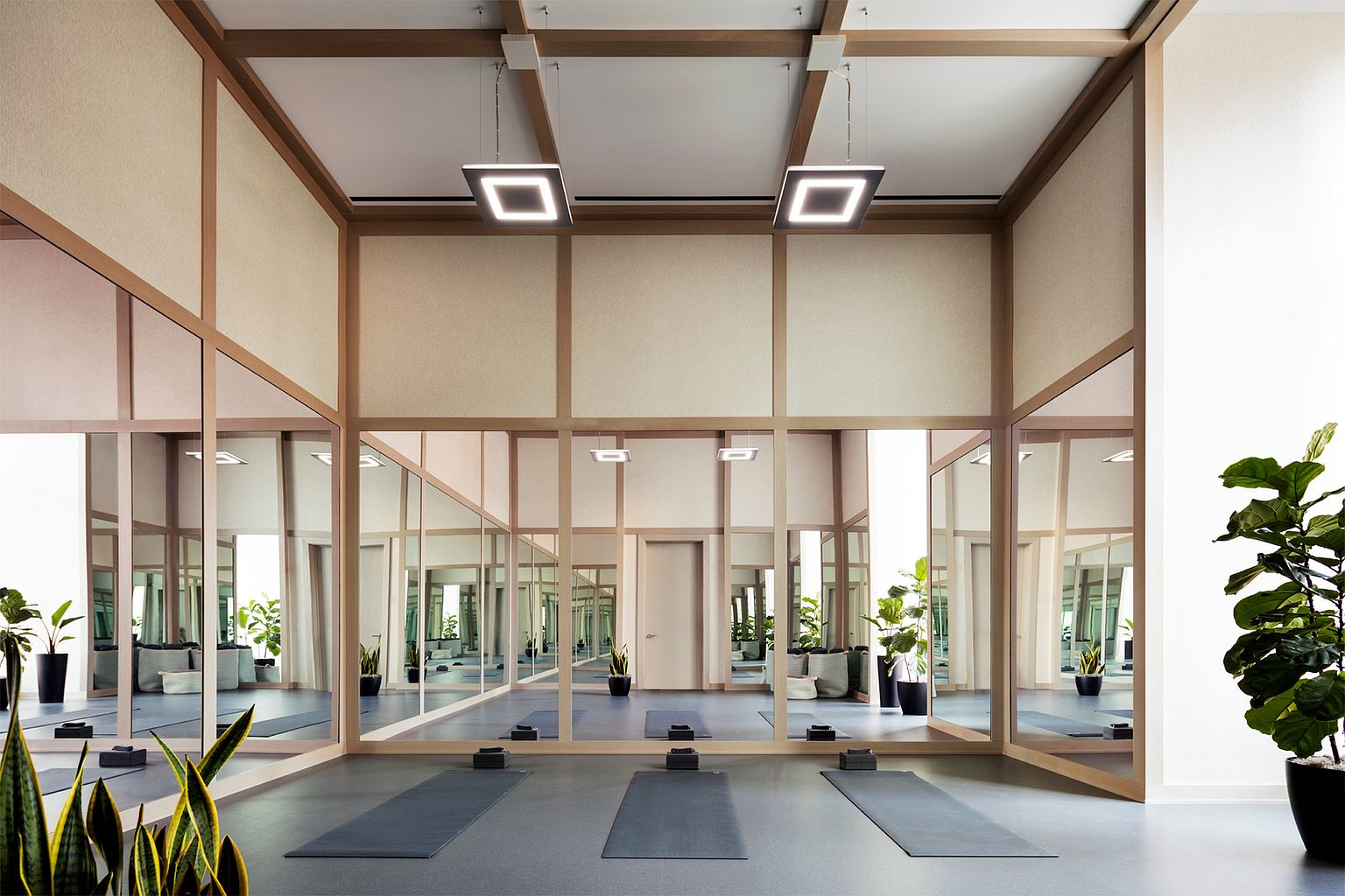 This serene yoga and fitness studio features a minimalist design with light wood framing and expansive mirrored walls that create a sense of depth and openness. Three dark yoga mats are neatly arranged on the smooth floor, accompanied by yoga blocks, while modern square pendant lights hang from the ceiling. A large potted plant adds a touch of greenery to the corner, enhancing the calm and professional atmosphere of the space.