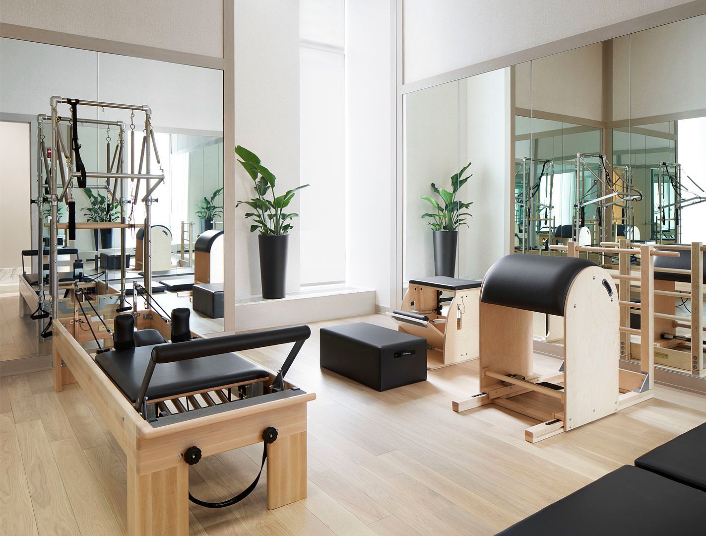 This bright and minimalist home gym is dedicated to Pilates, featuring professional-grade equipment including a reformer, a barrel, and a chair. The space is accented by light wood flooring, large mirrors that enhance the sense of depth, and two potted plants that add a touch of greenery. The clean, neutral aesthetic creates a serene and focused environment for exercise.