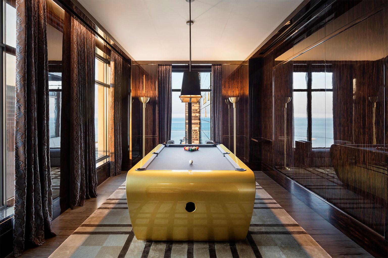 This sophisticated game room features a striking, gold-finished pool table as its centerpiece, set against a backdrop of rich, dark wood-paneled walls. Large windows on either side provide expansive views of the ocean and city skyline, while elegant floor-to-ceiling curtains frame the scenery. The room is illuminated by a modern pendant light and sleek floor lamps, creating a luxurious and cinematic atmosphere.