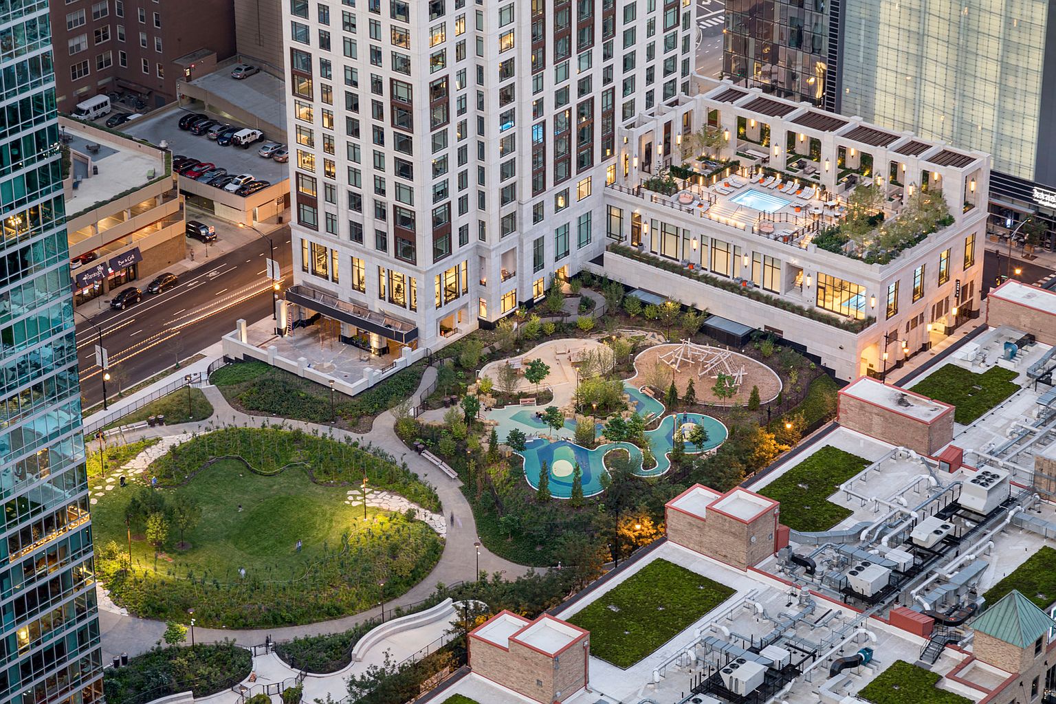 This high-angle aerial view captures a sophisticated urban residential complex featuring a lush, landscaped park and playground area nestled between modern high-rise buildings. The scene highlights a rooftop terrace complete with a swimming pool and lounge seating, offering a blend of luxury amenities and green space. The perspective emphasizes the integration of nature and architecture within a dense city environment, showcasing a well-maintained community setting.