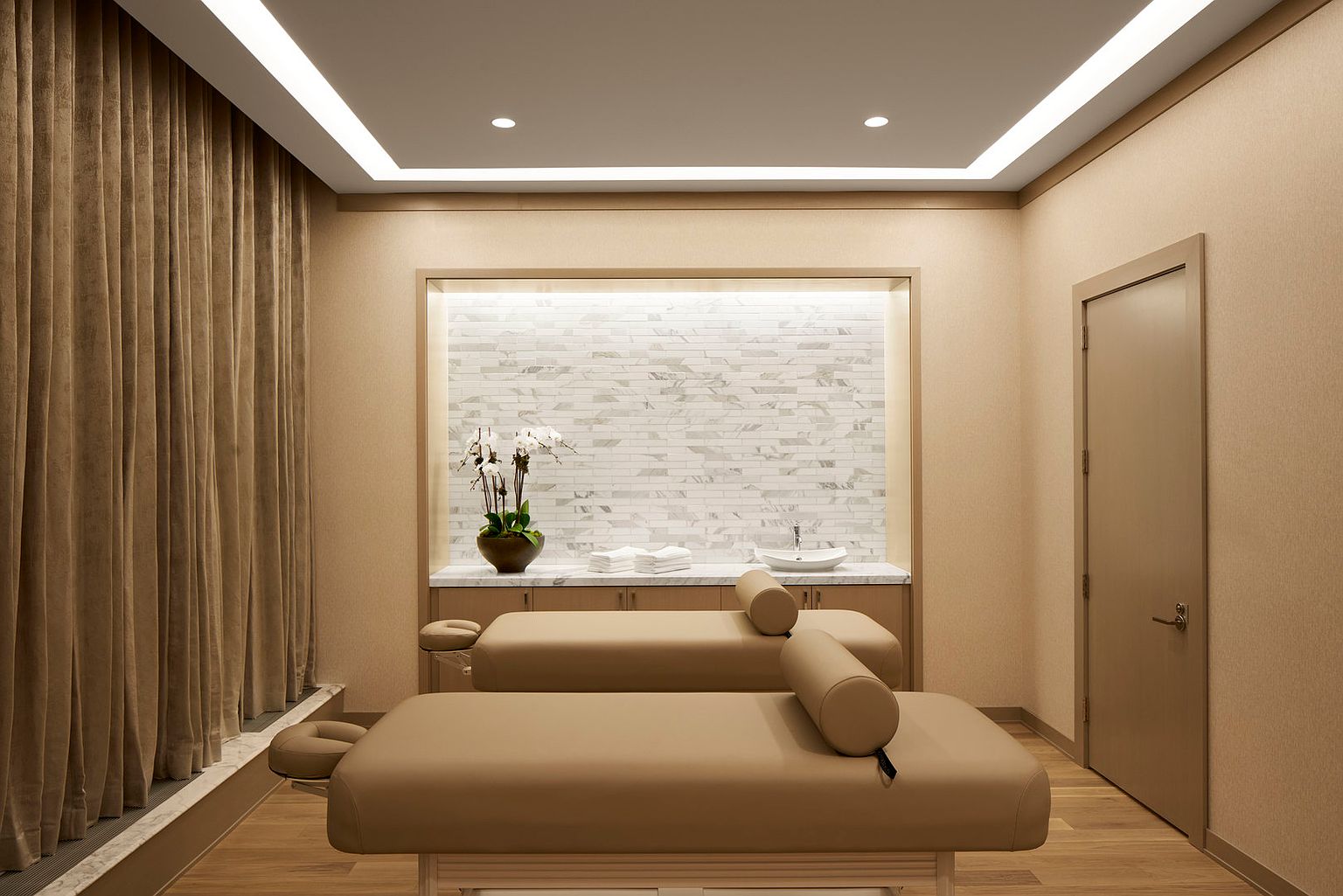 This professional spa or massage room features two treatment tables arranged in parallel, each equipped with a bolster pillow. The room is designed with a neutral color palette, including light wood flooring, beige walls, and floor-to-ceiling curtains along one side. A built-in vanity area at the back includes a stone countertop, a small sink, and a decorative stone-tiled wall, all illuminated by recessed lighting.