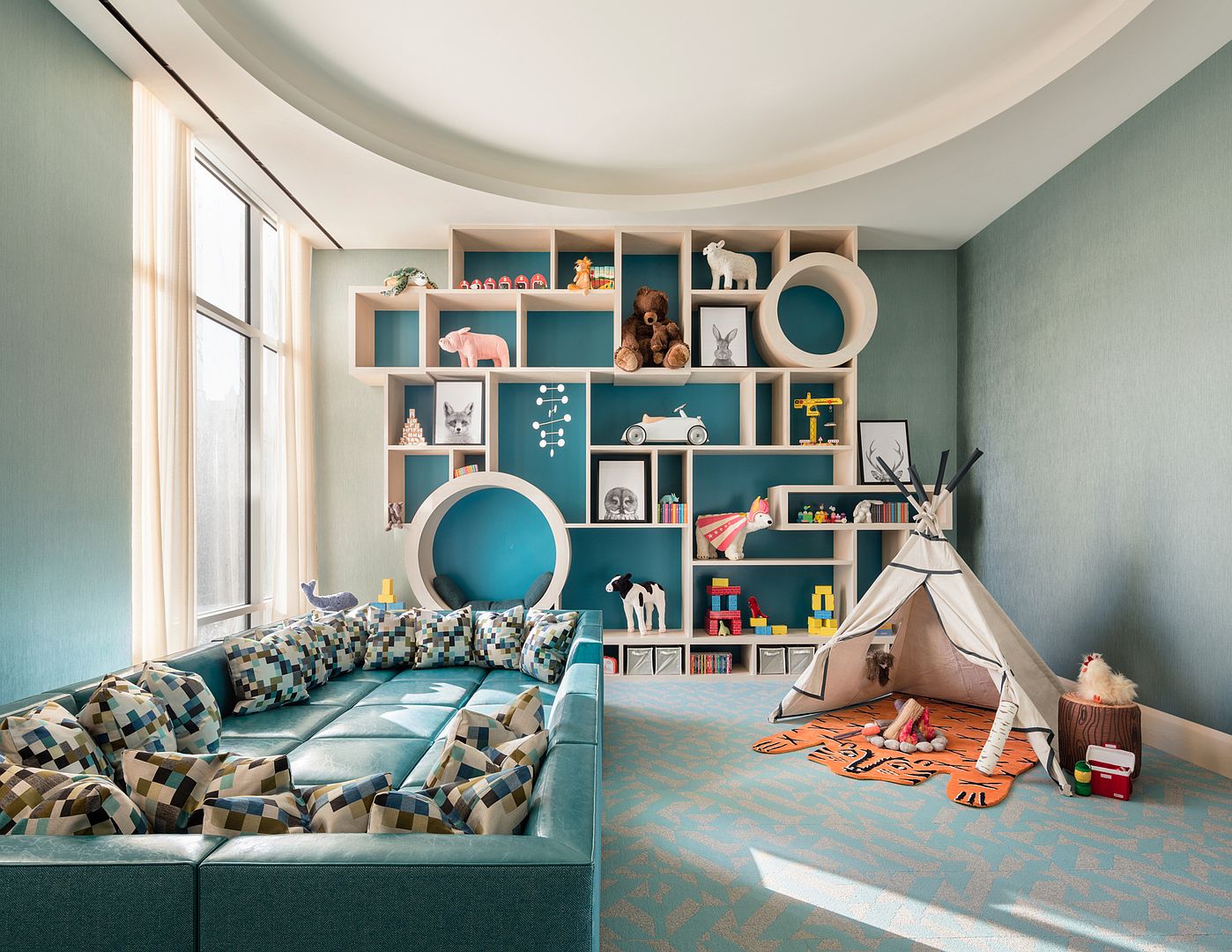 This vibrant children's playroom features a large, custom-built modular shelving unit that serves as a focal point, housing various toys, books, and decorative items. A deep, teal-colored sectional sofa with patterned cushions occupies the foreground, while a whimsical teepee tent sits on a tiger-patterned rug in the corner. The room is bathed in natural light from tall windows, creating a playful and inviting atmosphere with a cohesive blue and light wood color palette.