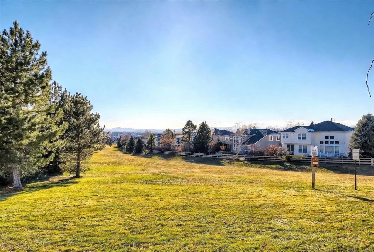 This wide-angle shot captures a sprawling, grassy backyard that transitions into a scenic golf course or open green space. In the background, several large, multi-story suburban homes are lined up behind a wooden fence, offering a sense of community and privacy. The bright, clear blue sky and mature trees framing the left side create a peaceful, expansive atmosphere ideal for outdoor living.