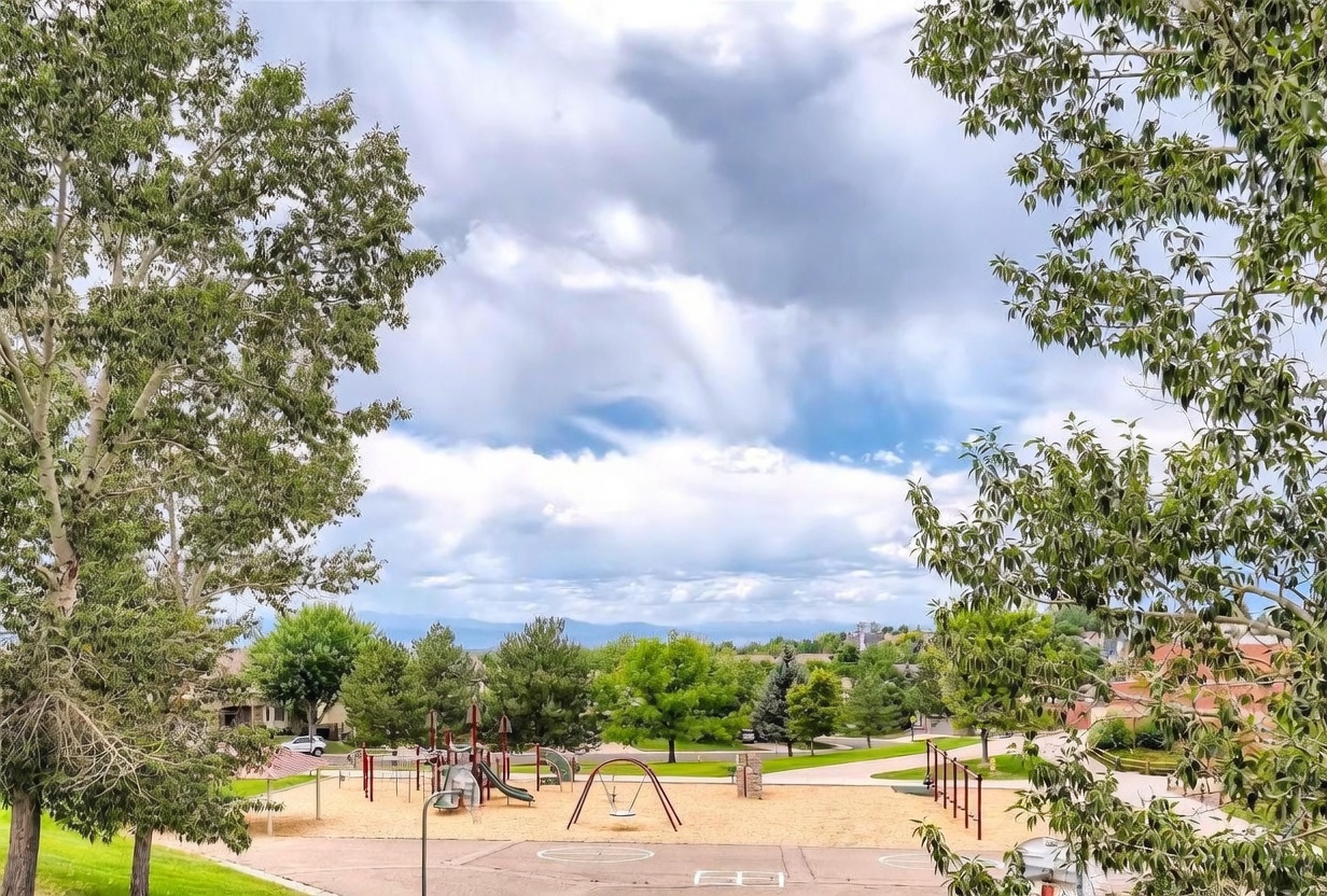 This elevated, wide-angle shot captures a vibrant community park featuring a playground with swings, slides, and climbing structures set against a backdrop of lush green trees and distant mountains. The foreground includes a paved area, likely a basketball court, leading into the sandy play zone under a dramatic, cloud-filled sky. The perspective provides a scenic, panoramic view that highlights the recreational amenities available within the neighborhood.