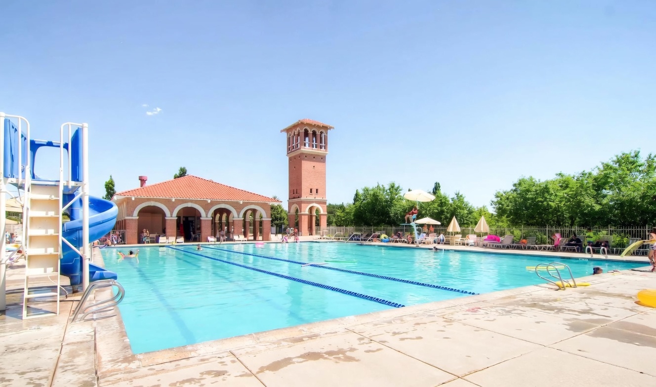 This image showcases a large, inviting community swimming pool featuring a blue spiral water slide and a lifeguard chair. In the background, a distinctive brick building with a tall, tower-like architectural feature adds a unique aesthetic to the recreational area. The scene is captured from a ground-level perspective, emphasizing the expansive pool deck and the bright, sunny atmosphere perfect for community leisure.