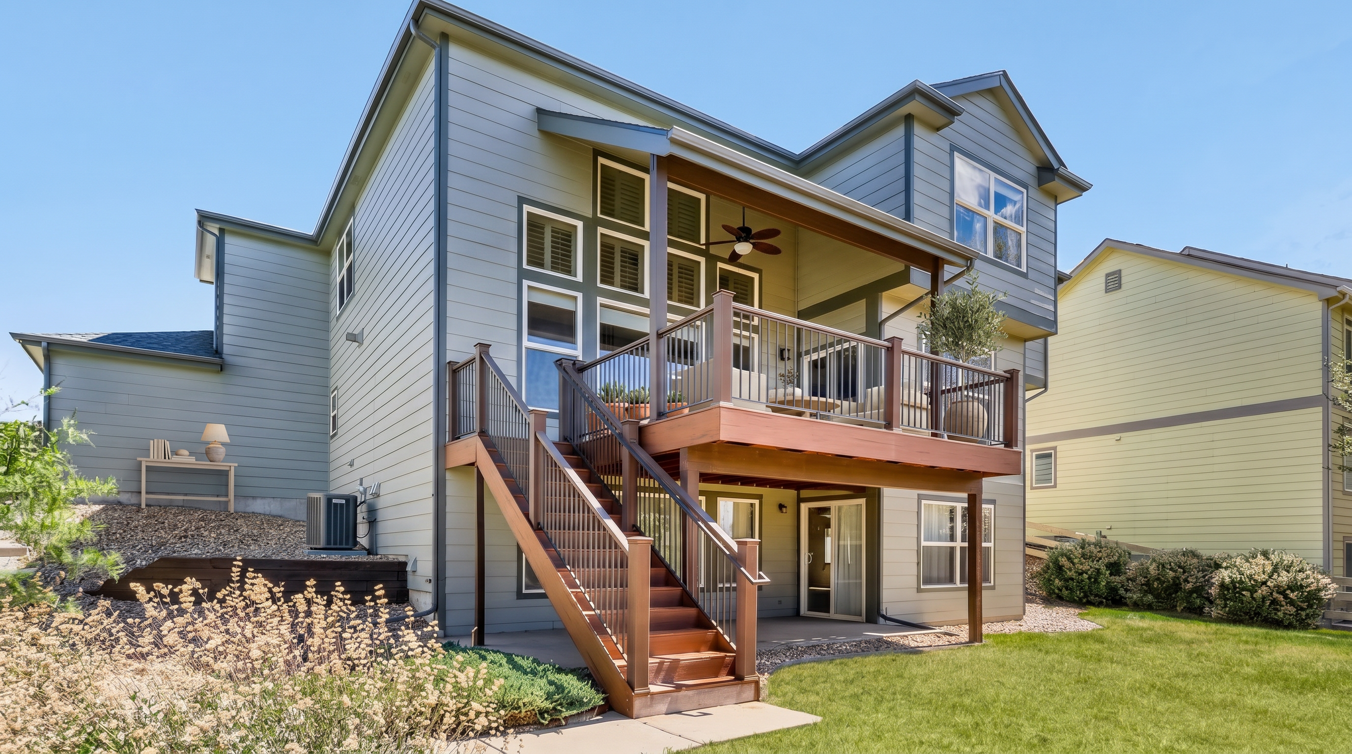 This rear view showcases a multi-level house with light grey horizontal siding and a prominent, elevated wood deck featuring black metal railings and a staircase leading down to the patio. The home includes a series of tall, stacked windows and a covered porch area with a ceiling fan, providing a great space for outdoor living. The backyard is landscaped with a mix of rock beds, decorative bushes, and a neat lawn, bordering a neighboring yellow-toned home.