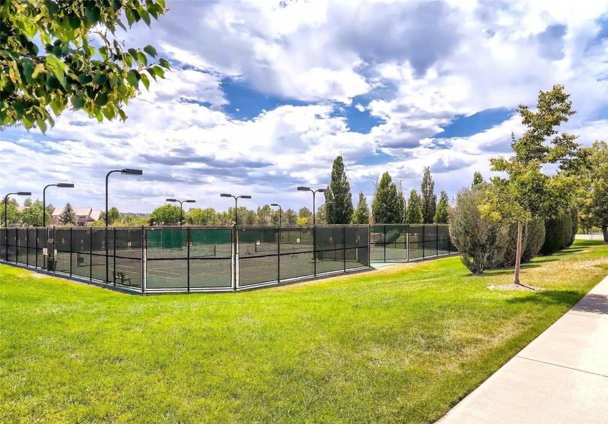 This image showcases a well-maintained community tennis court facility enclosed by black chain-link fencing. The scene is set against a backdrop of lush green grass, mature trees, and a vibrant blue sky with scattered clouds, creating an inviting recreational atmosphere. The perspective is a wide-angle ground-level shot that emphasizes the openness and accessibility of the neighborhood amenity.