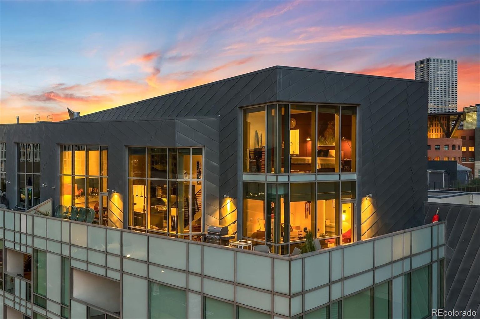 This aerial view showcases a modern, multi-story building with a distinctive gray exterior and large windows, offering a glimpse into the well-lit interiors. The building features a spacious balcony with outdoor furniture and a grill, suggesting a luxurious urban living experience. The backdrop includes a vibrant sunset sky and a cityscape, enhancing the property's appeal.