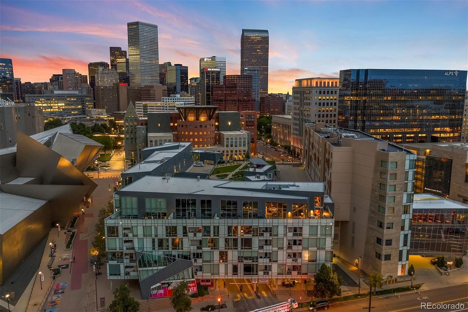 This aerial view showcases a modern building with a unique architectural design, featuring a mix of glass and concrete elements. The building is situated in a vibrant urban setting, surrounded by other high-rise buildings and city infrastructure. The image captures the building's exterior during a beautiful sunset, highlighting the city skyline.