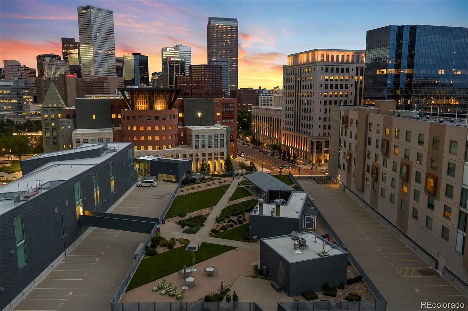 This aerial view showcases a modern urban landscape at dusk, featuring a mix of residential and commercial buildings. The foreground highlights a well-maintained courtyard with green spaces and seating areas, while the background presents a stunning skyline with illuminated skyscrapers against a colorful sky. The overall impression is one of sophisticated city living with access to outdoor amenities.