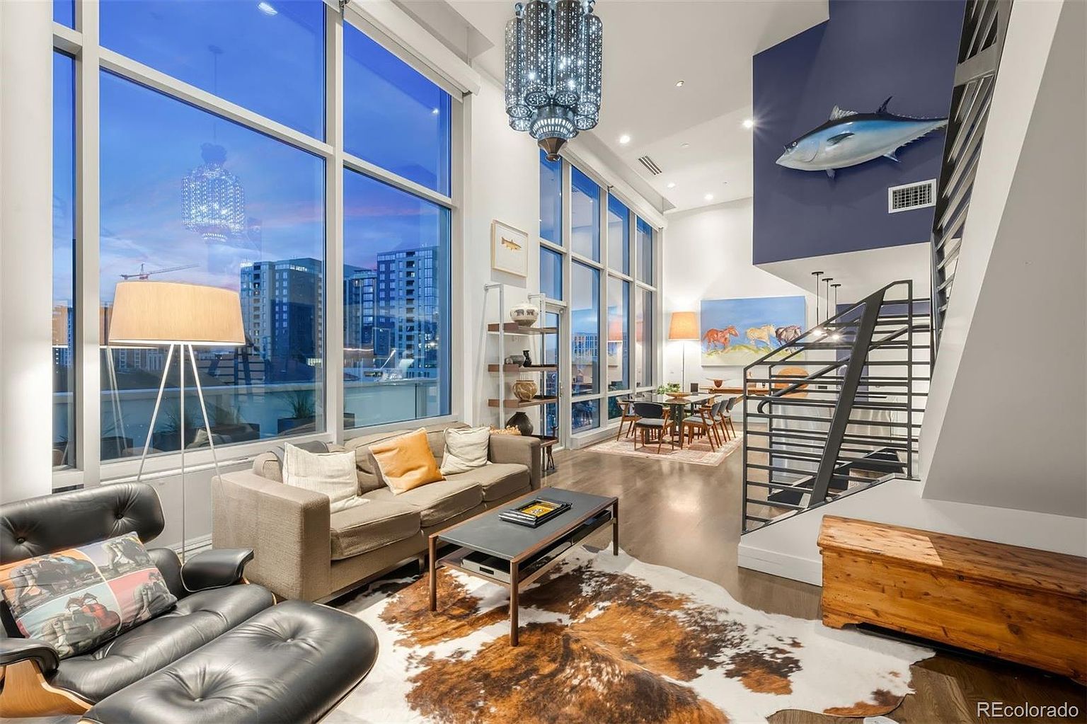 This is a spacious and modern living room with floor-to-ceiling windows offering city views. The room features a comfortable sofa, a leather lounge chair, a unique cowhide rug, and a stylish coffee table. A staircase leads to another level, and artwork adds character to the space, creating an inviting and sophisticated atmosphere.