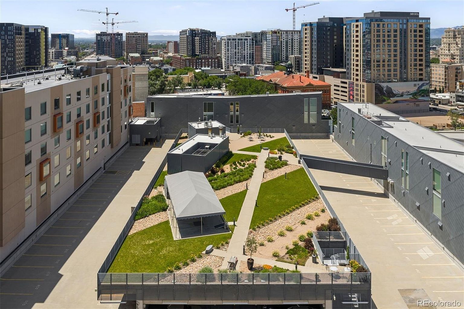 This aerial view showcases a modern building complex with a unique rooftop garden and parking area. The rooftop garden features green spaces, walkways, and a covered structure, offering a recreational amenity. The surrounding cityscape includes other high-rise buildings and construction cranes, indicating an urban environment.