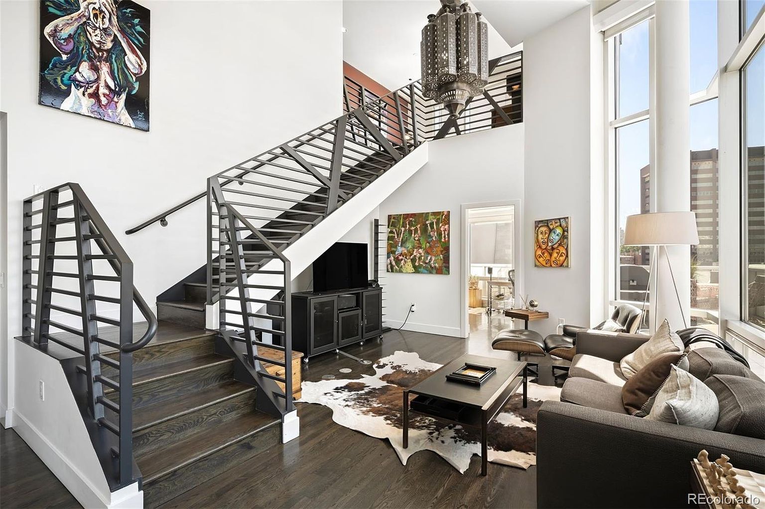 This interior shot showcases a modern living room with a striking staircase featuring metal railings. The room is decorated with contemporary art and a cowhide rug, adding a touch of texture. Natural light floods the space through large windows, highlighting the dark hardwood floors and the comfortable-looking gray sofa.
