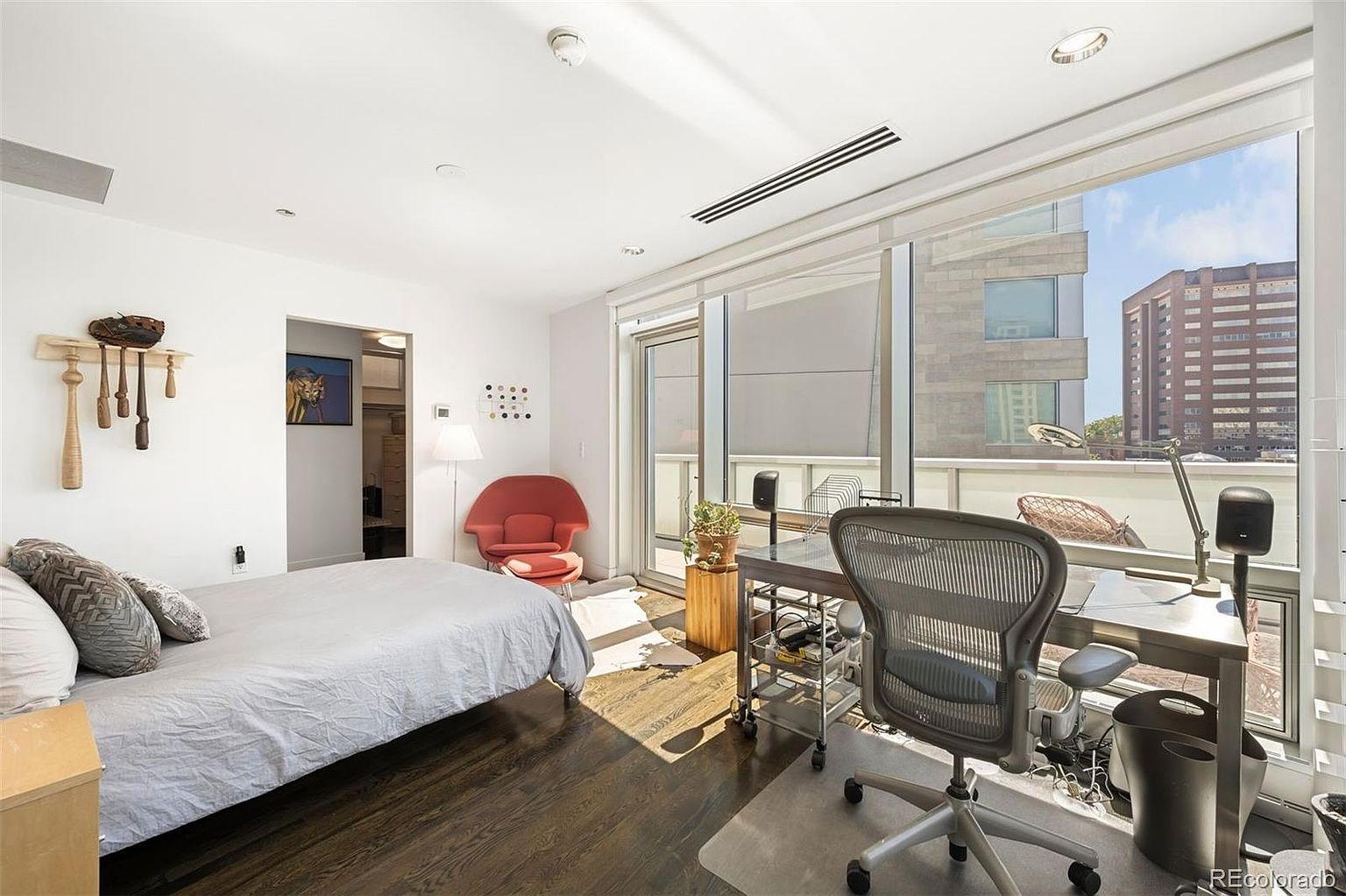 This primary bedroom features a modern design with a bed, a desk area by a large window, and hardwood floors. The room is well-lit with natural light and includes a red accent chair and unique wall decor. The overall impression is a comfortable and stylish living space.
