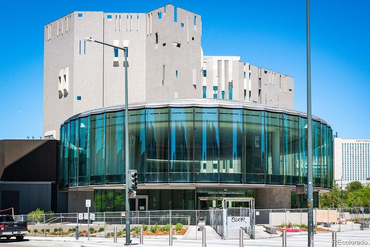 This is a front exterior view of a modern building with a unique architectural design. The building features a cylindrical glass structure attached to a larger, block-like structure with a light-colored facade. The entrance is visible, and the surrounding area includes landscaping and street elements, suggesting a commercial or public building in an urban setting.