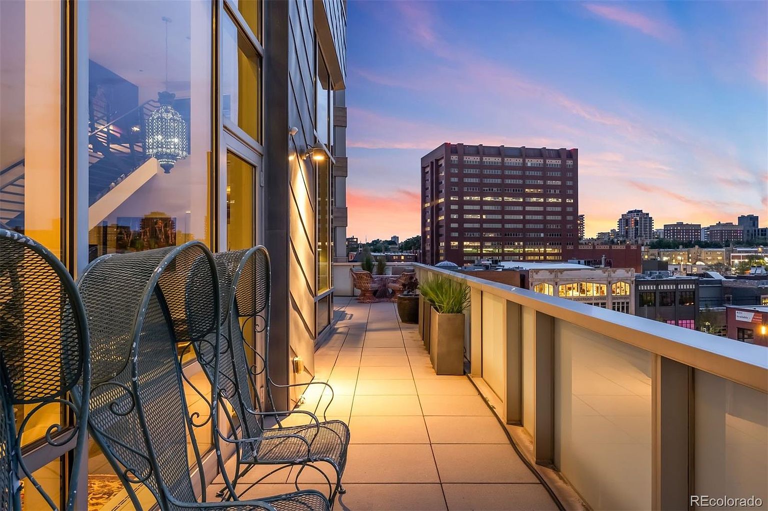 This image showcases a stylish balcony with metal chairs, offering a stunning cityscape view at dusk. The balcony features a modern glass railing and tiled flooring, creating an inviting outdoor living space. The warm lighting and architectural details enhance the overall appeal, making it a desirable feature for potential buyers.