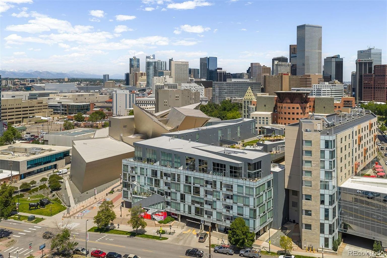 This aerial view showcases a modern building complex in an urban setting, with a backdrop of a city skyline and distant mountains. The architecture features a mix of glass, concrete, and metal elements, creating a contemporary aesthetic. The surrounding area includes green spaces and streetscapes, adding to the property's appeal.