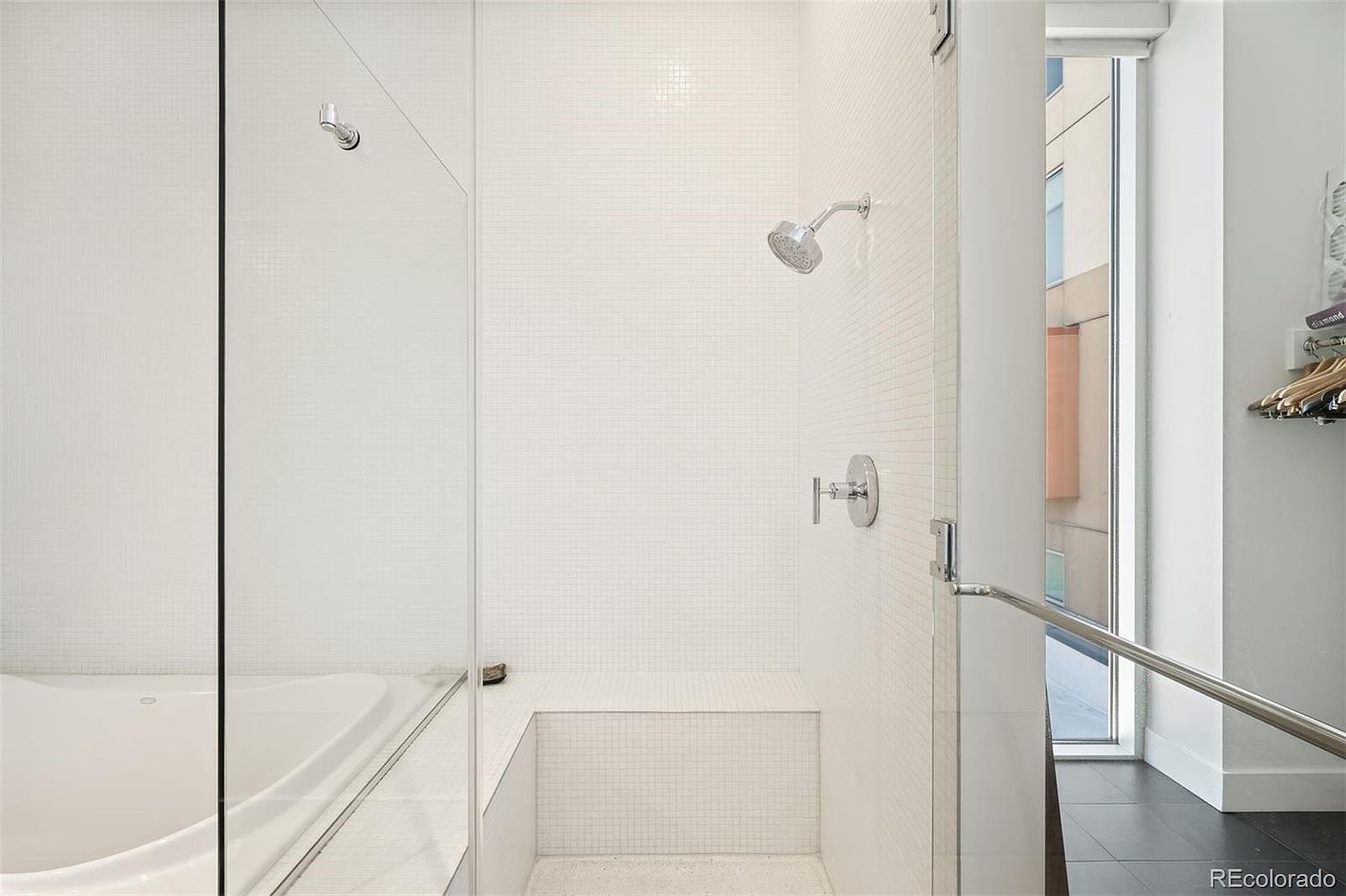 This is a bright and modern primary bathroom featuring a walk-in shower with white mosaic tile walls and a built-in bench. The shower has a glass door and chrome fixtures, including a shower head and handle. Natural light enters from a nearby window, creating a clean and spa-like atmosphere.