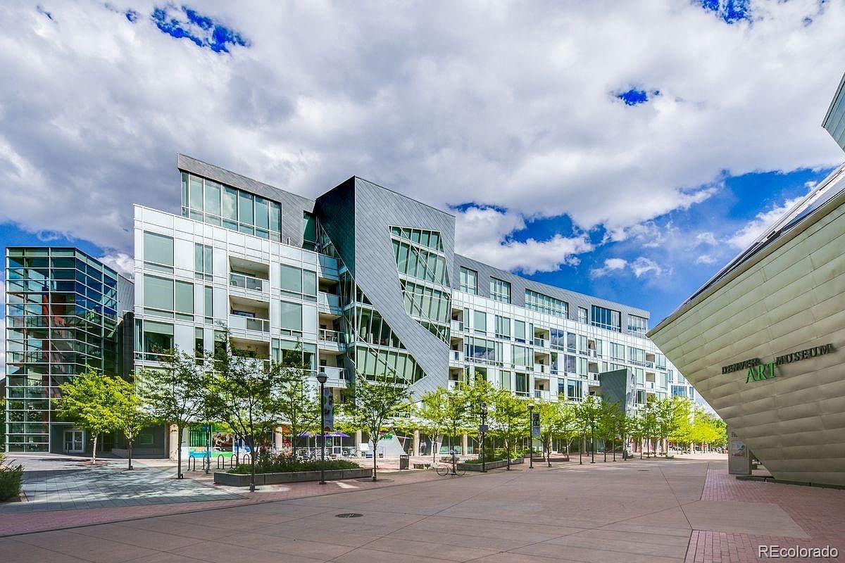 This image showcases the front view of a modern architectural building complex, possibly a museum or residential building, under a partly cloudy sky. The building features a mix of glass and metal facade, with a unique angular design. The surrounding area includes a paved plaza with trees, creating a welcoming and aesthetically pleasing environment.
