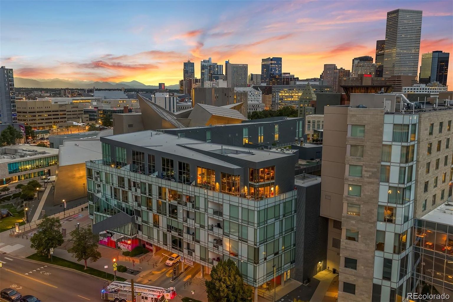 This aerial view showcases a modern building complex against a stunning sunset backdrop with city skyline and distant mountains. The building features a mix of glass and stone facade, with visible balconies and well-lit interiors. The overall impression is one of urban sophistication and luxurious living.